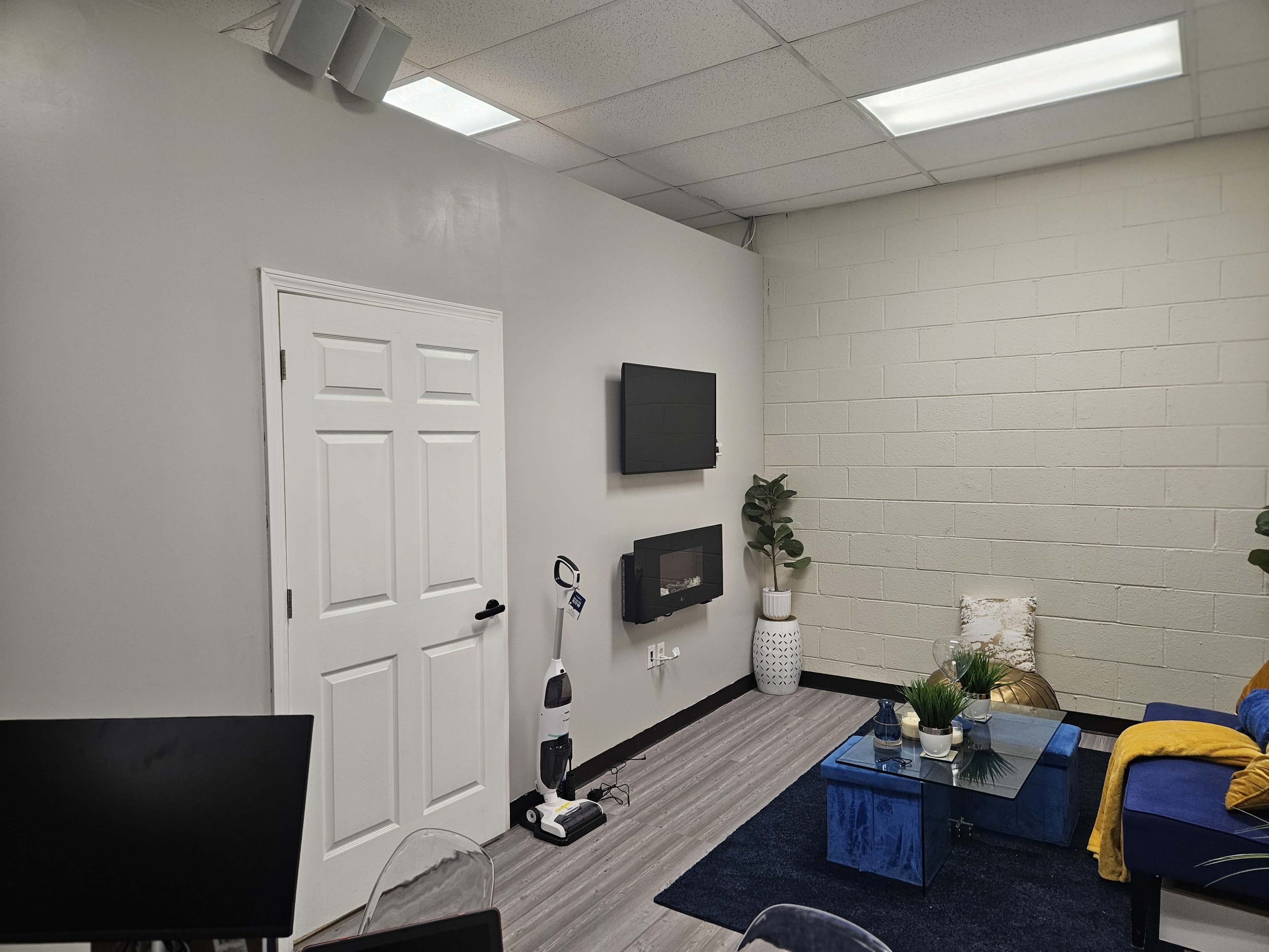 The image shows a modern office space featuring a wall-mounted television, a door, a vacuum cleaner on the floor, and a blue coffee table with decorative plants.