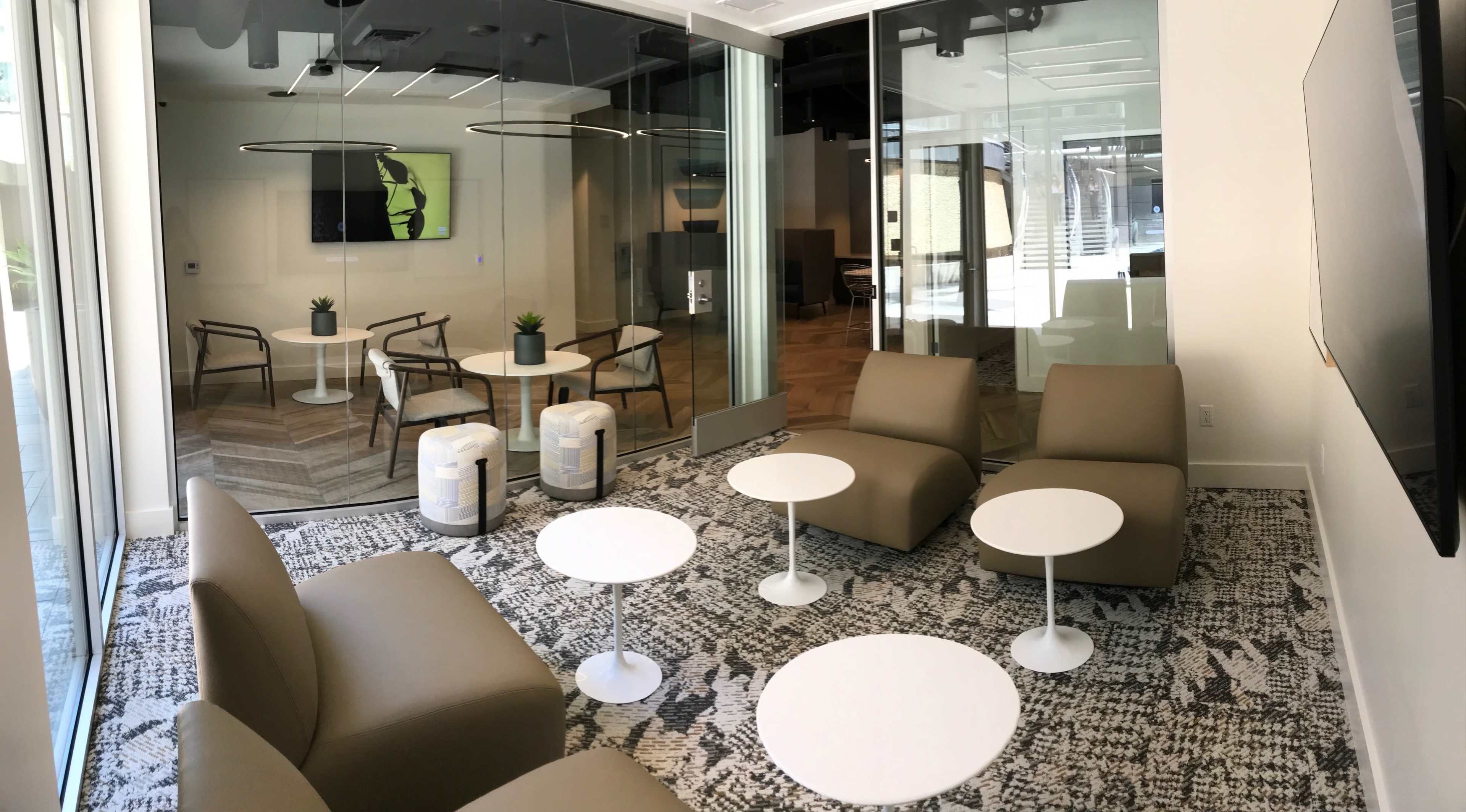 A modern lounge area featuring low seating and small white tables, with a large glass wall separating it from another space.