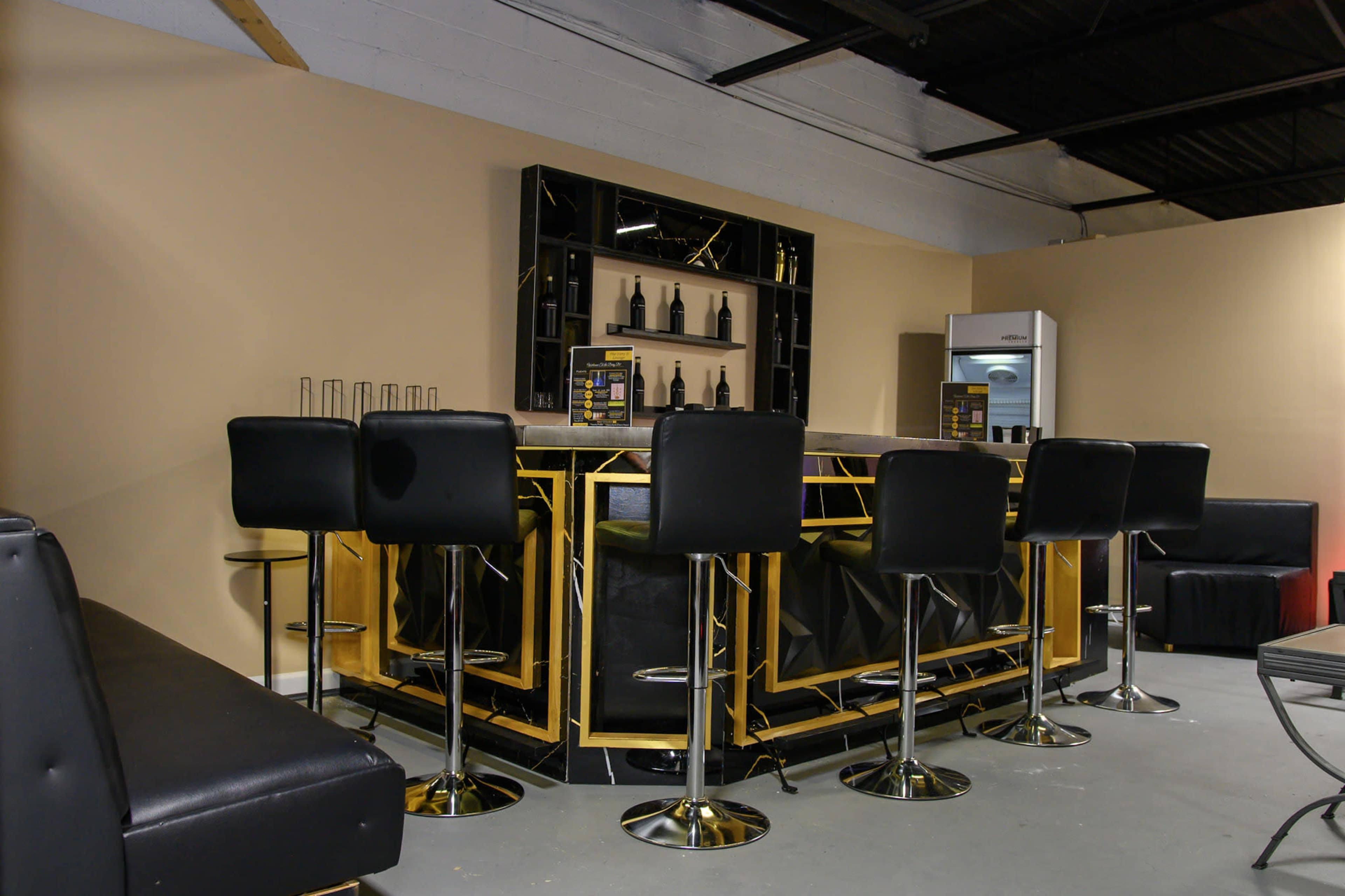 The image shows a modern bar area with a glossy black and gold countertop, high-backed stools, and a shelf displaying bottles in a minimalist setting.