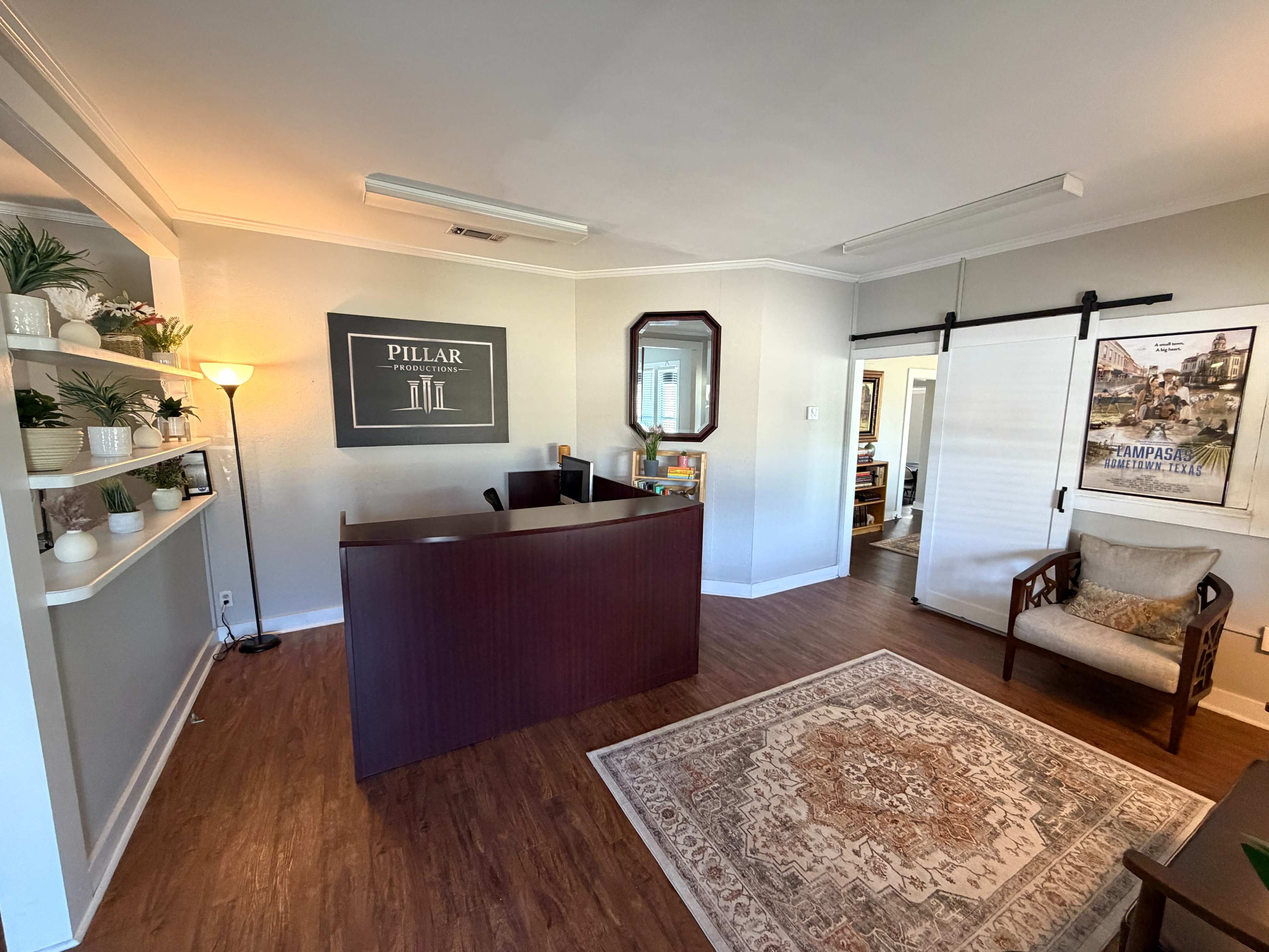The image shows a tidy office reception area featuring a wooden front desk, a decorative rug, a large mirror, and plants on a shelf.