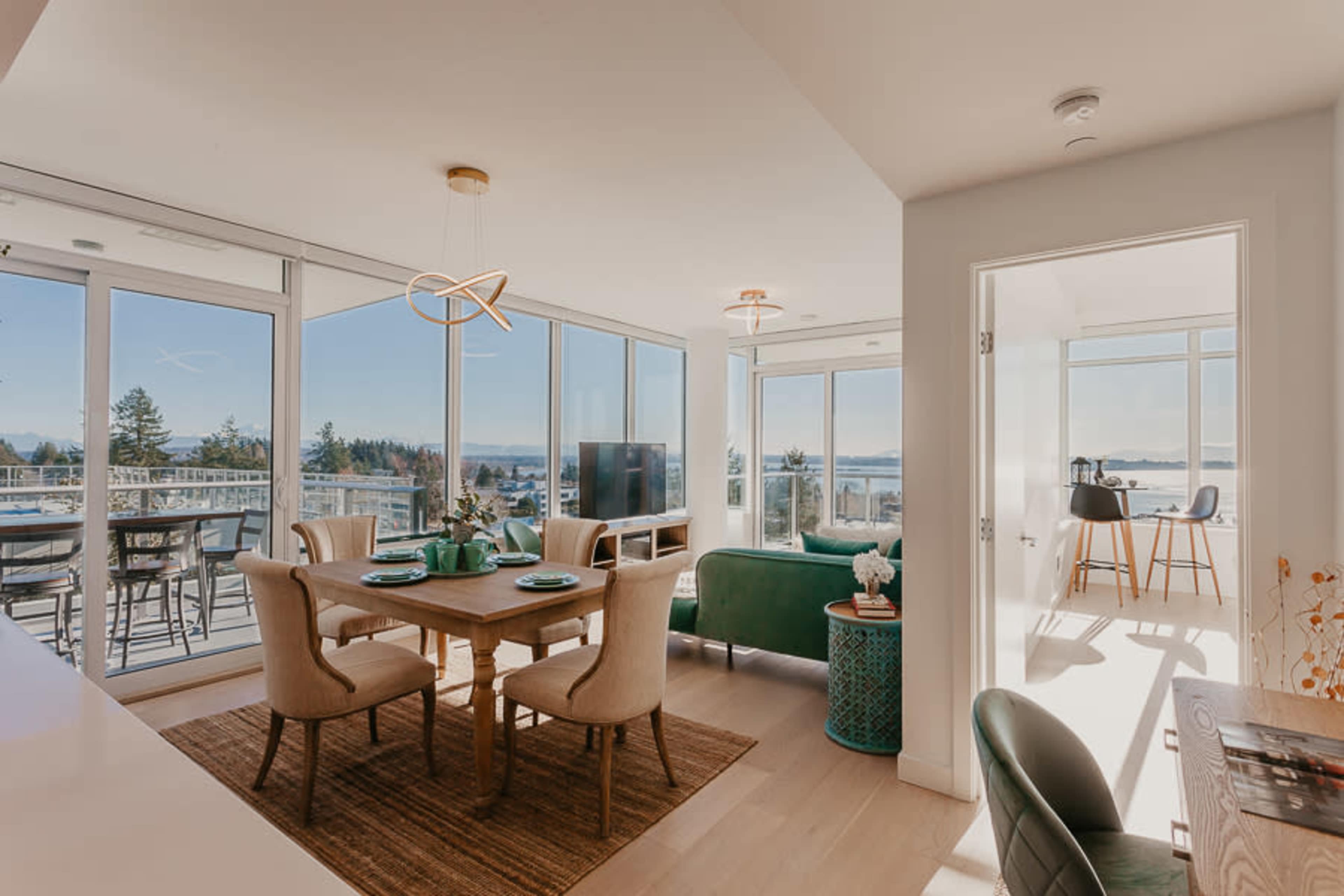 A modern open-concept living and dining area with large windows offering views of the outdoors, a dining table set for four, and a cozy seating area with a green couch.