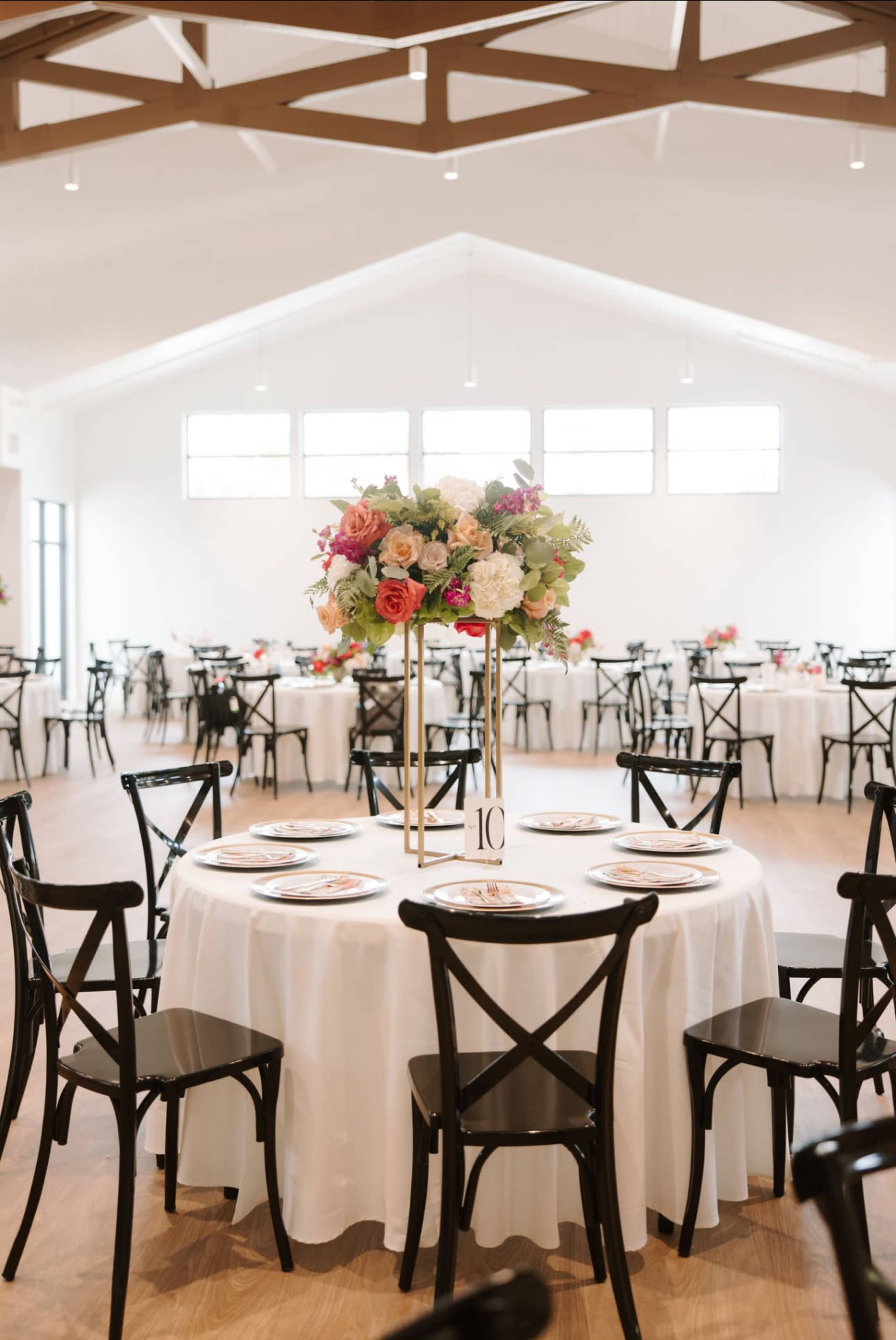 A spacious dining area features round tables with floral centerpieces and black wooden chairs, illuminated by natural light from large windows.