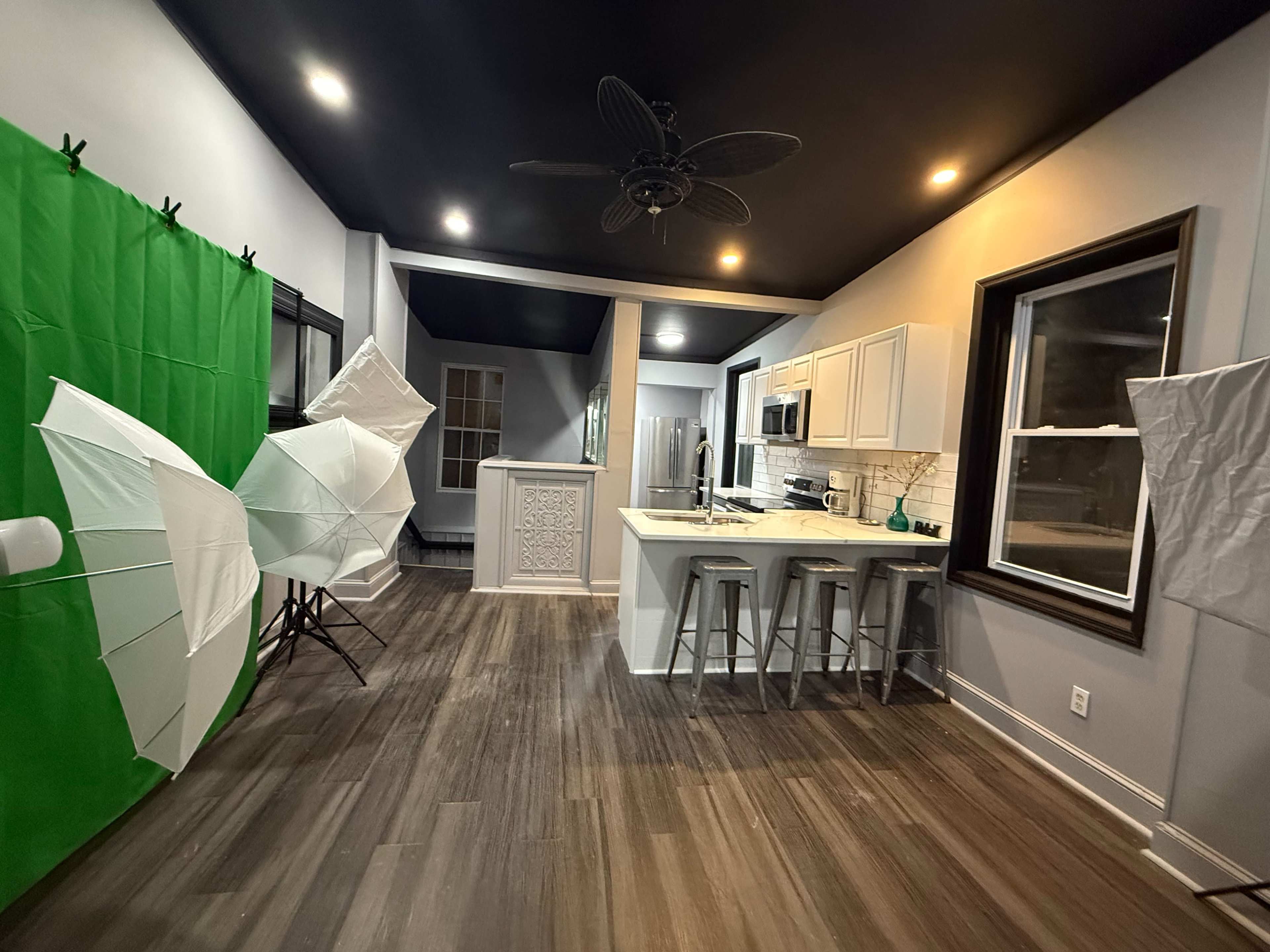 The image shows a spacious kitchen with white cabinets, a countertop, and a green backdrop set up for photography, alongside several light umbrellas.