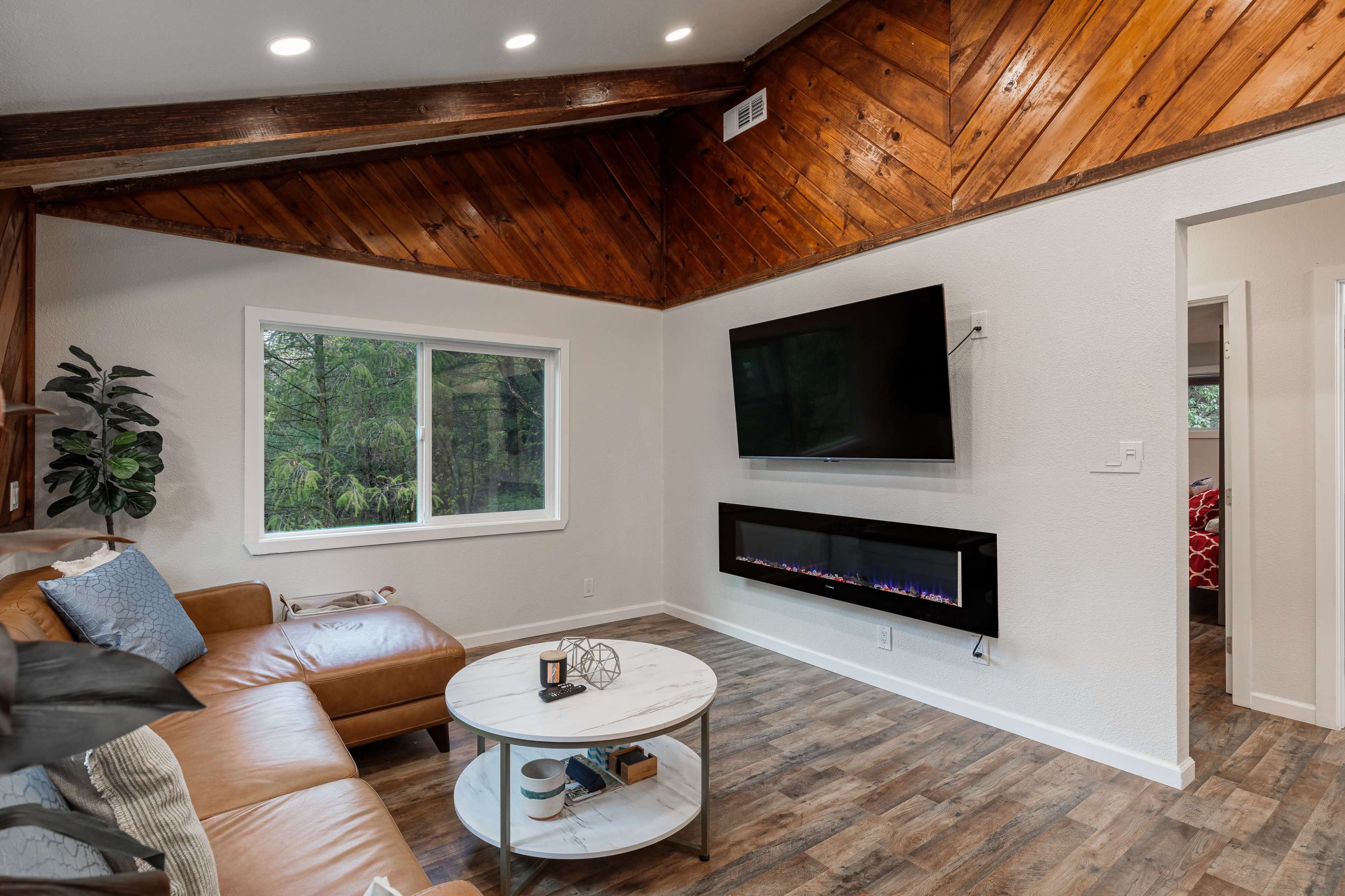 A cozy living room with a brown leather couch, a marble-top coffee table, a wall-mounted TV, and an electric fireplace beneath it, all set against a backdrop of wooden paneling and a large window overlooking greenery.