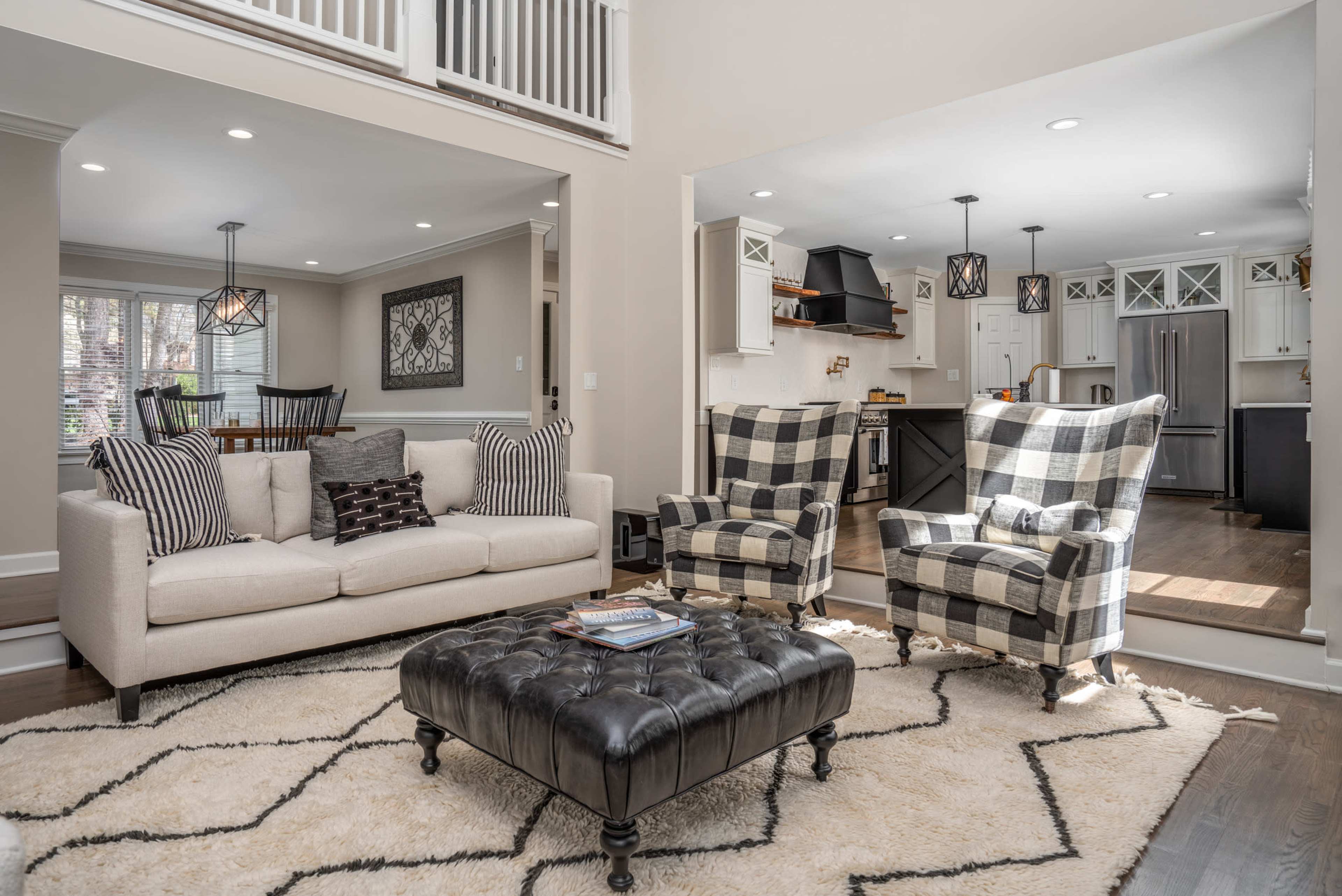 A living room with a beige sofa and two plaid armchairs positioned around a black ottoman on a light-colored rug, with an open view into a nearby kitchen.