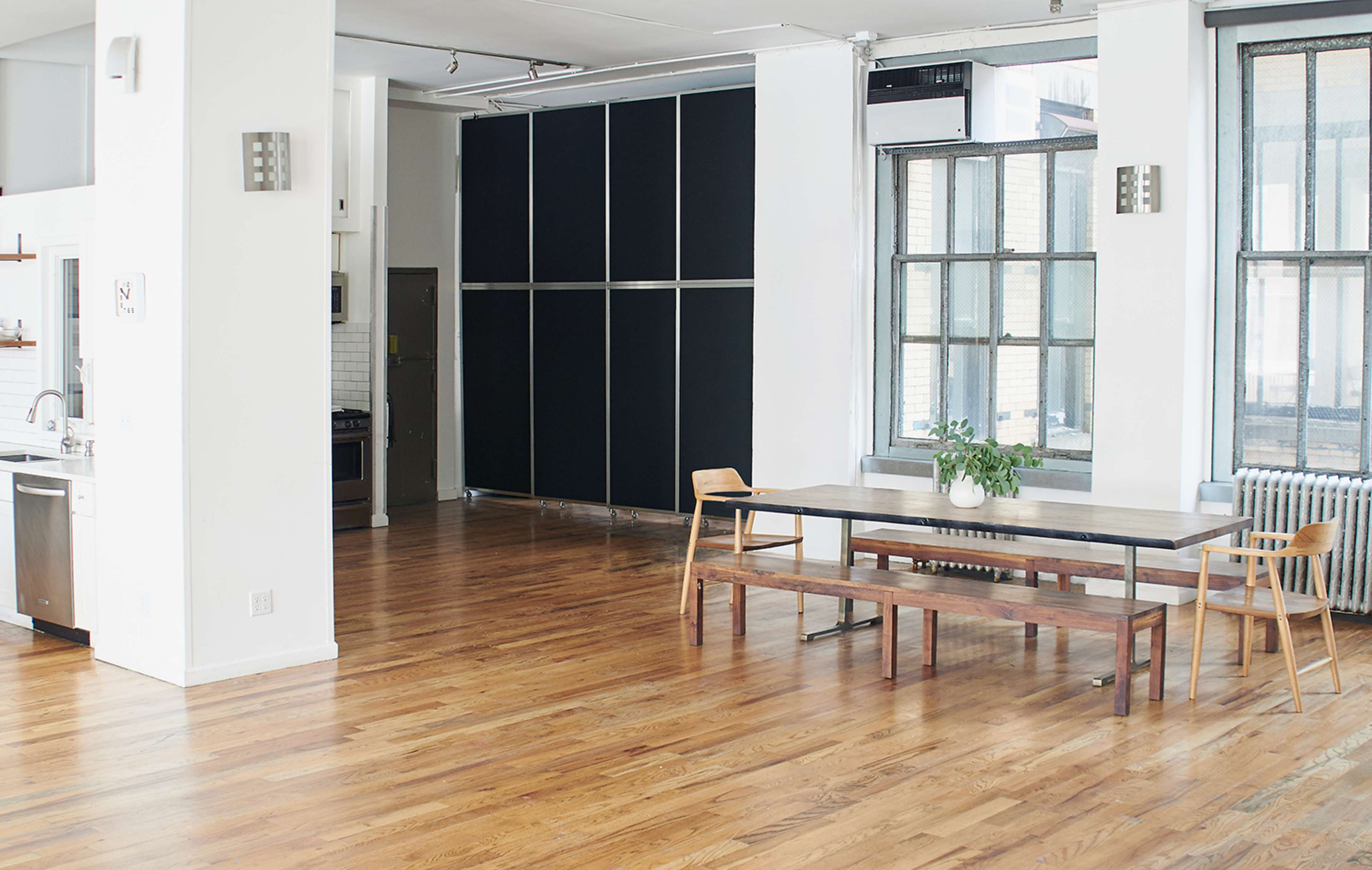 The image shows an open-plan, modern living space with wooden floors, a large dining table, and a kitchen area featuring stainless steel appliances.
