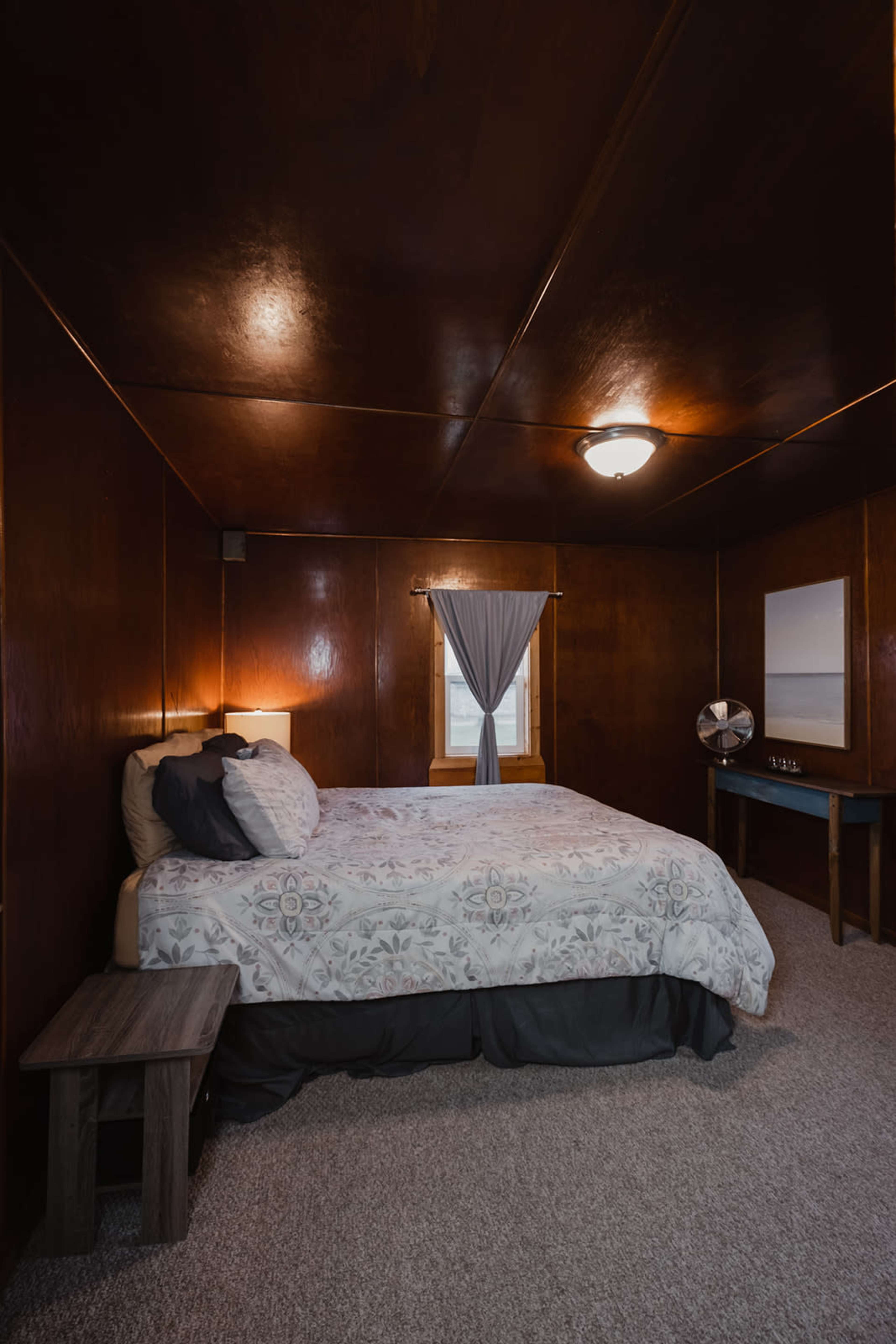 A simple bedroom with a double bed, a nightstand, a window with a grey curtain, and a small table with a mirror.