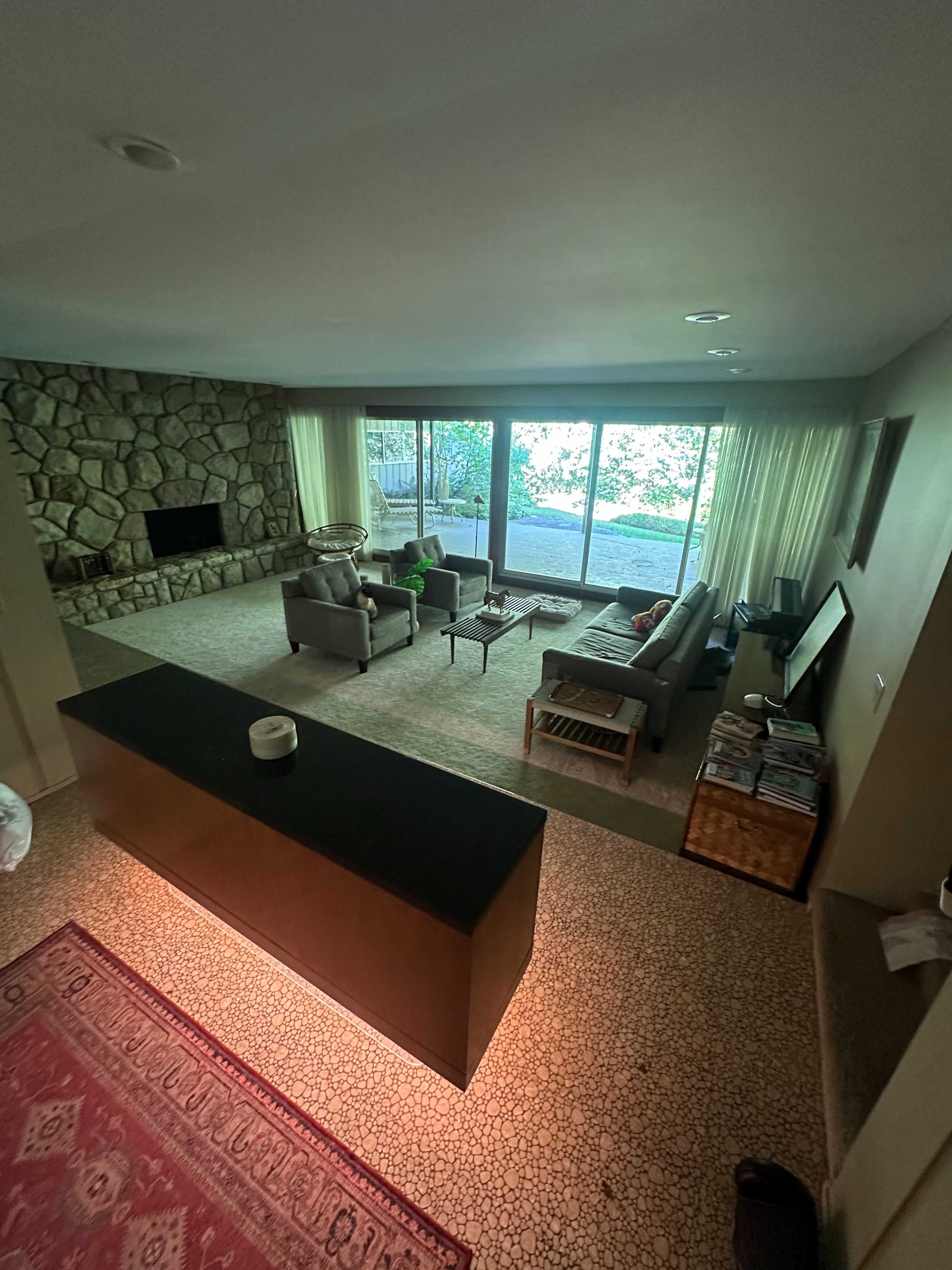 The image shows a spacious living room with stone walls, large windows letting in natural light, and a variety of furniture arranged around a low coffee table.
