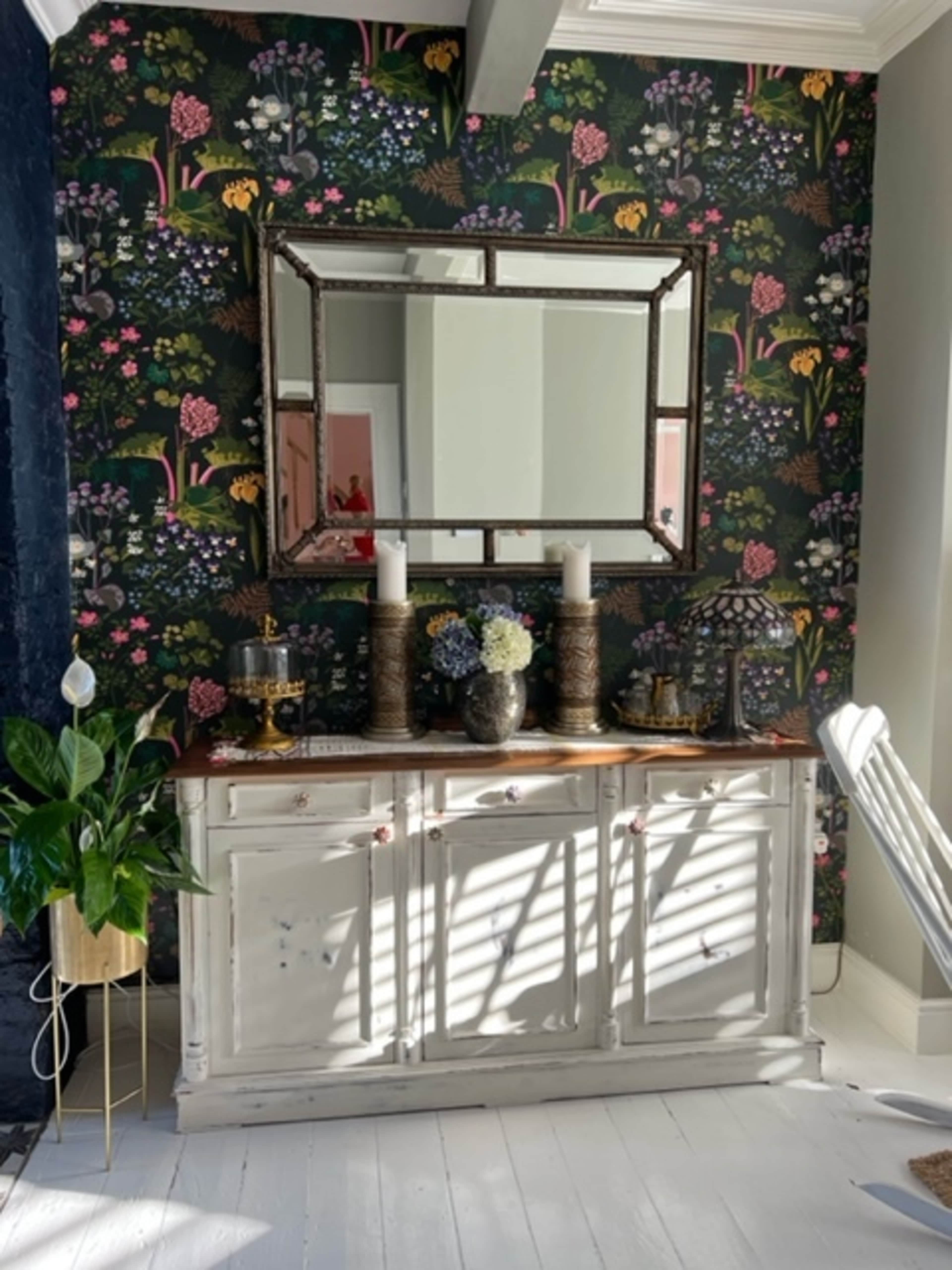 A white sideboard with decorative items and candles stands against a floral patterned wallpaper, reflecting a large mirror above it.