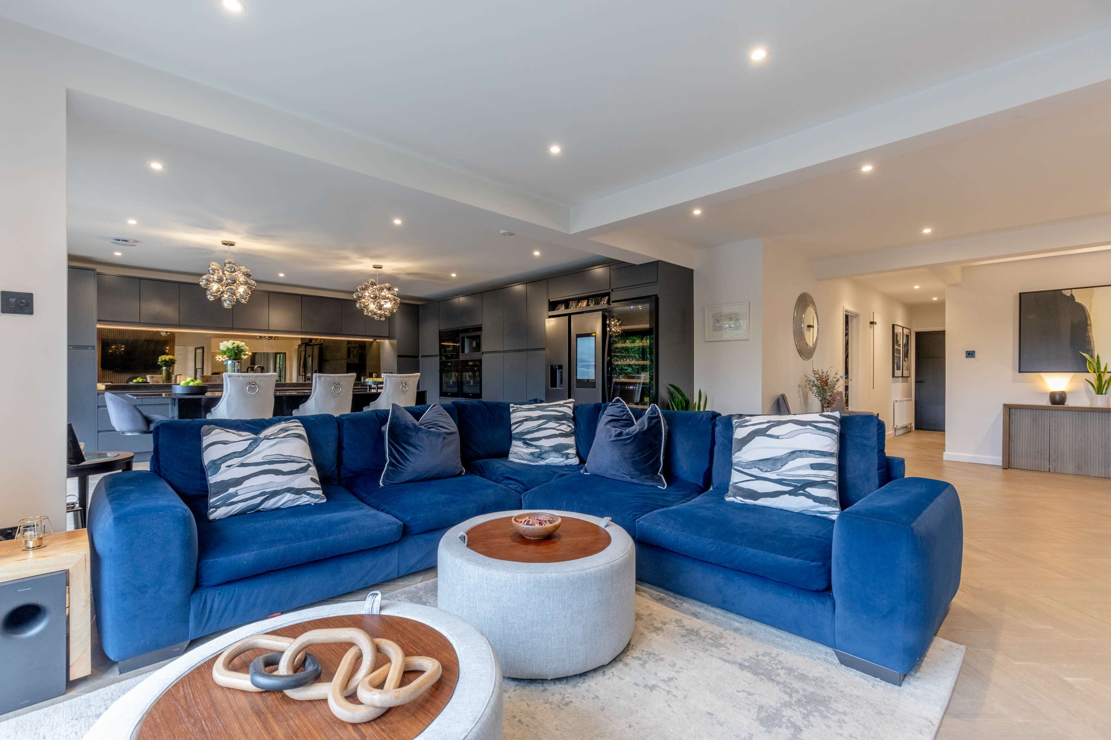 The image shows a modern living room with a large blue sectional sofa, a round coffee table, and a spacious kitchen in the background.