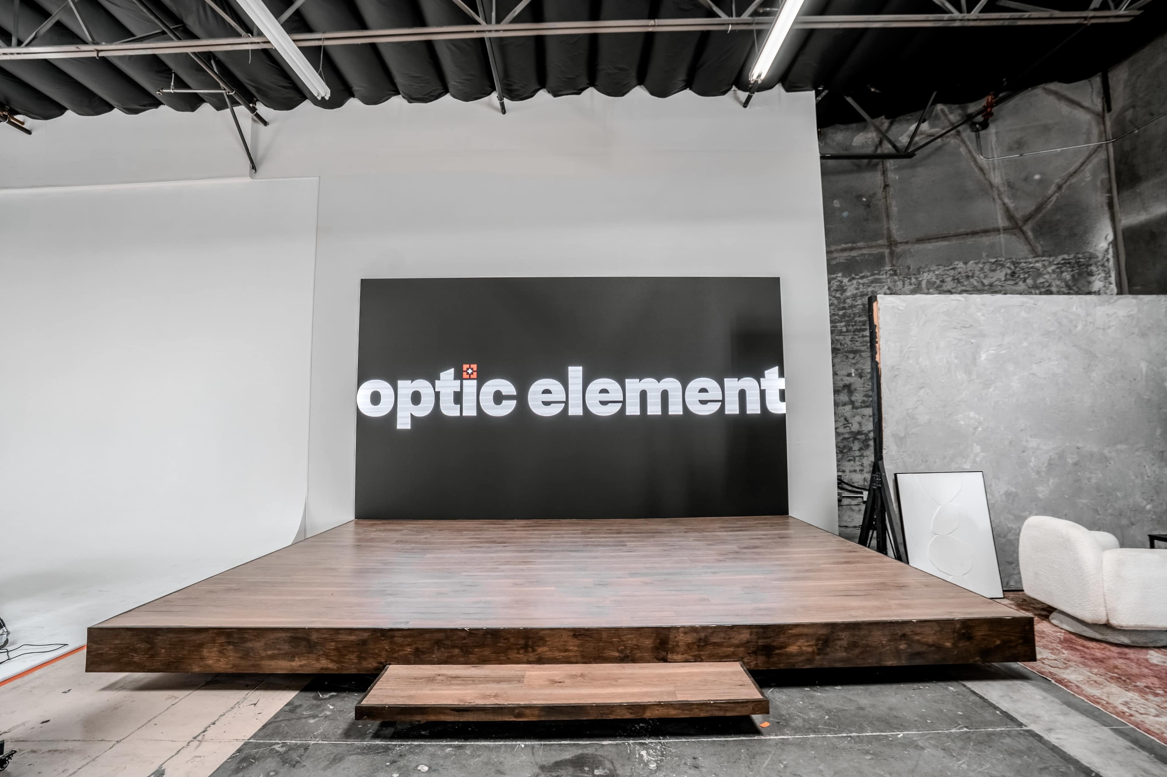 The image shows a stage setup with a large black backdrop displaying the text "optic element" in white, on a wooden surface with an empty backdrop on one side.