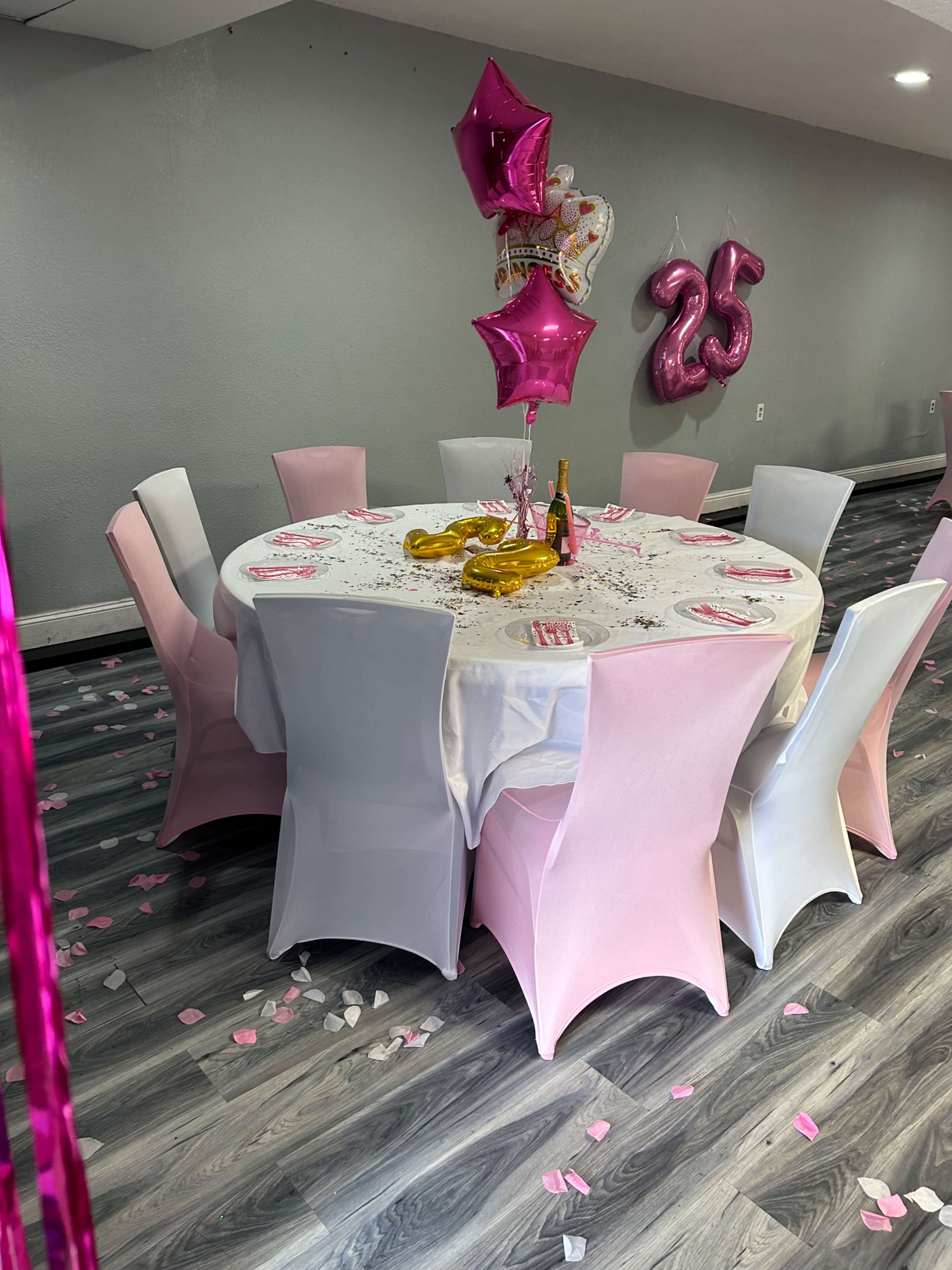 A round table is set with a white tablecloth and pink decorations, surrounded by chairs, with pink star-shaped balloons and a "25" balloon in the background.