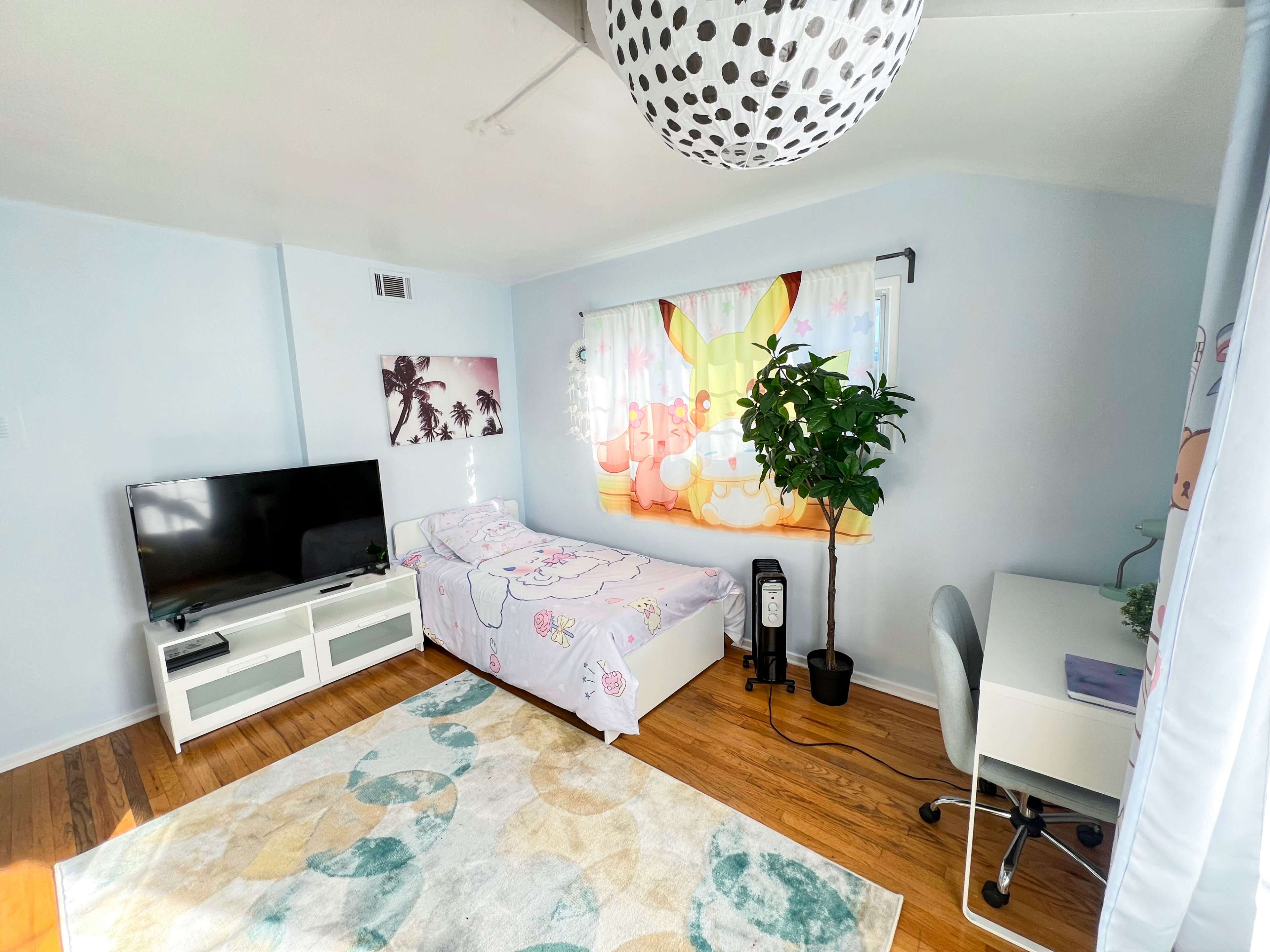The image shows a brightly lit bedroom featuring a single bed with a floral comforter, a television on a white stand, a desk with a chair, and a large plant near the window.