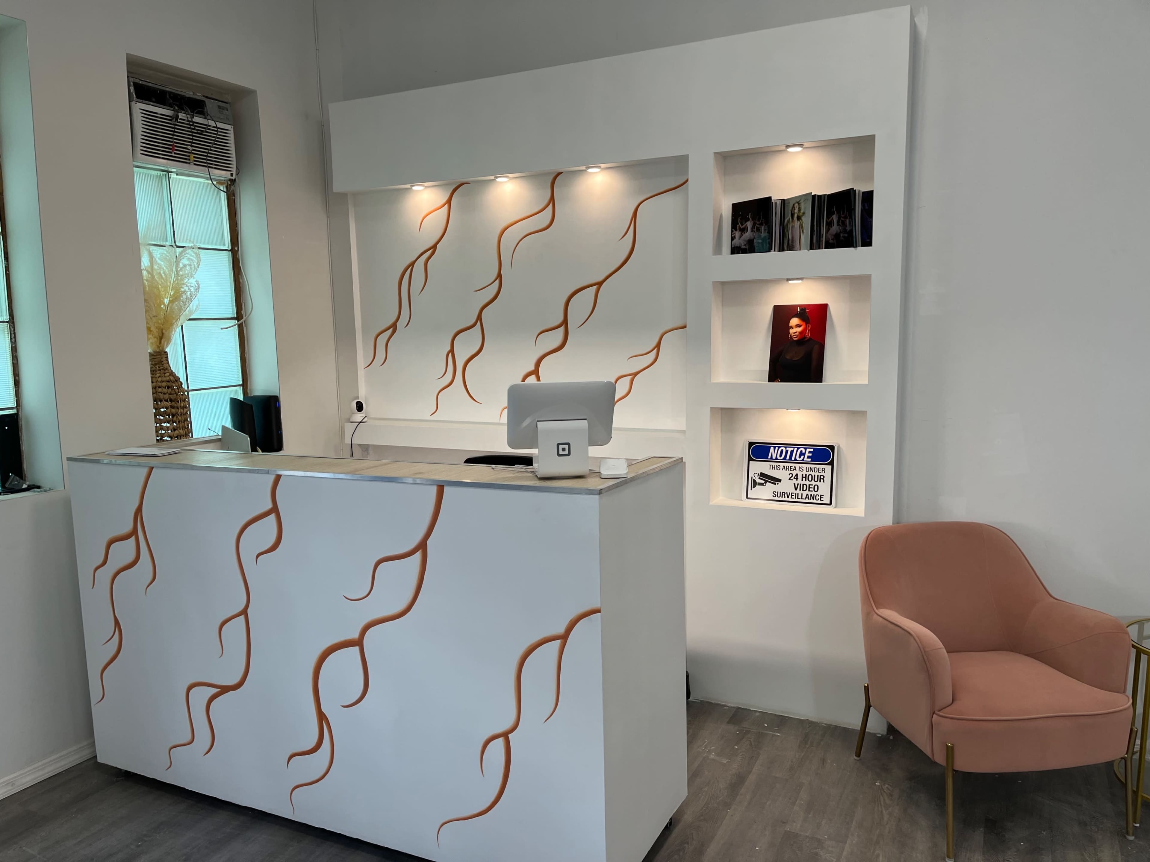 A sleek reception area with a white desk featuring decorative orange lines, illuminated shelving with framed photos, and a pink armchair beside it.