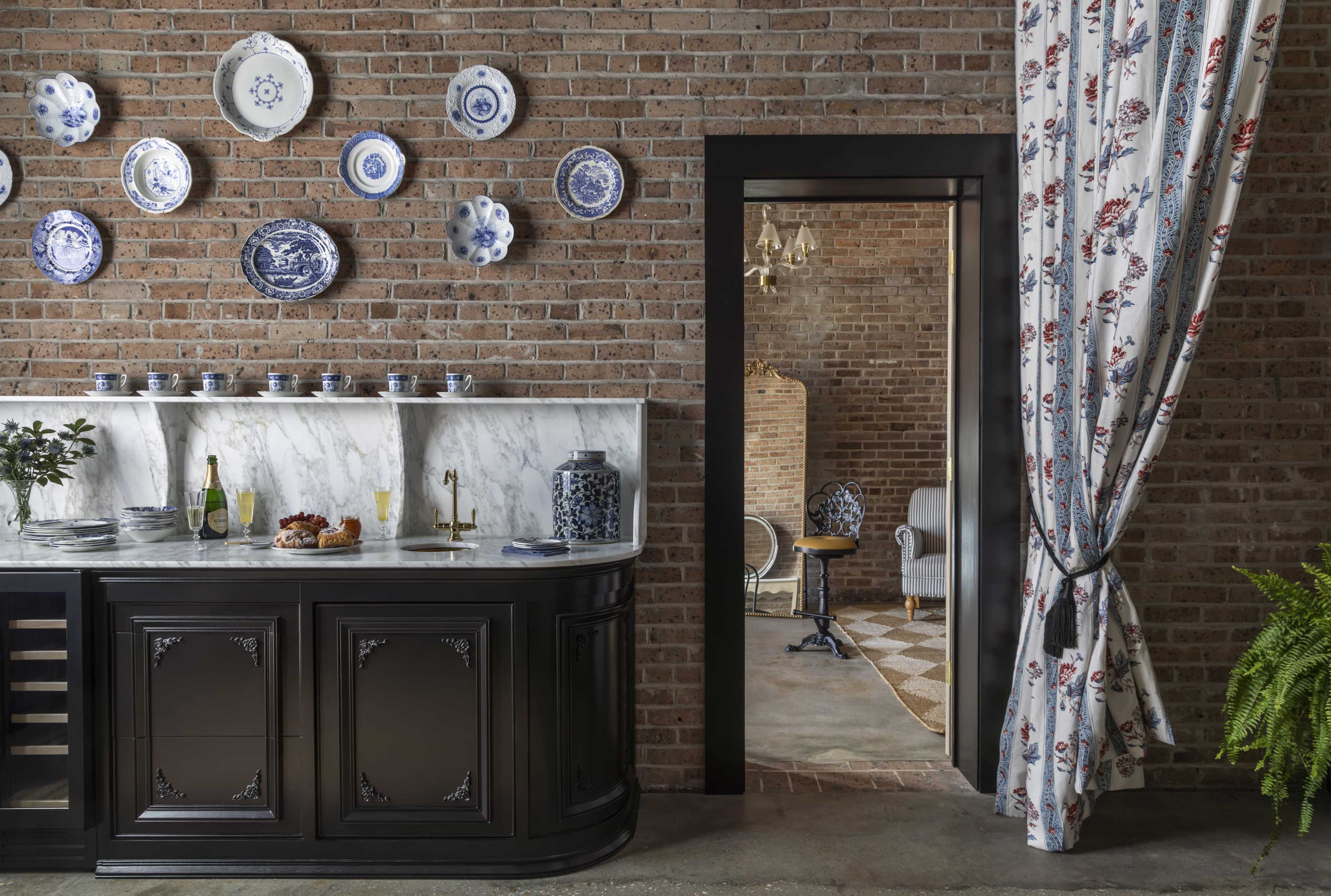 The image shows a modern kitchen with a black cabinetry, marble countertop, decorative blue and white plates on a brick wall, and a doorway leading to another room with a cozy seating area.