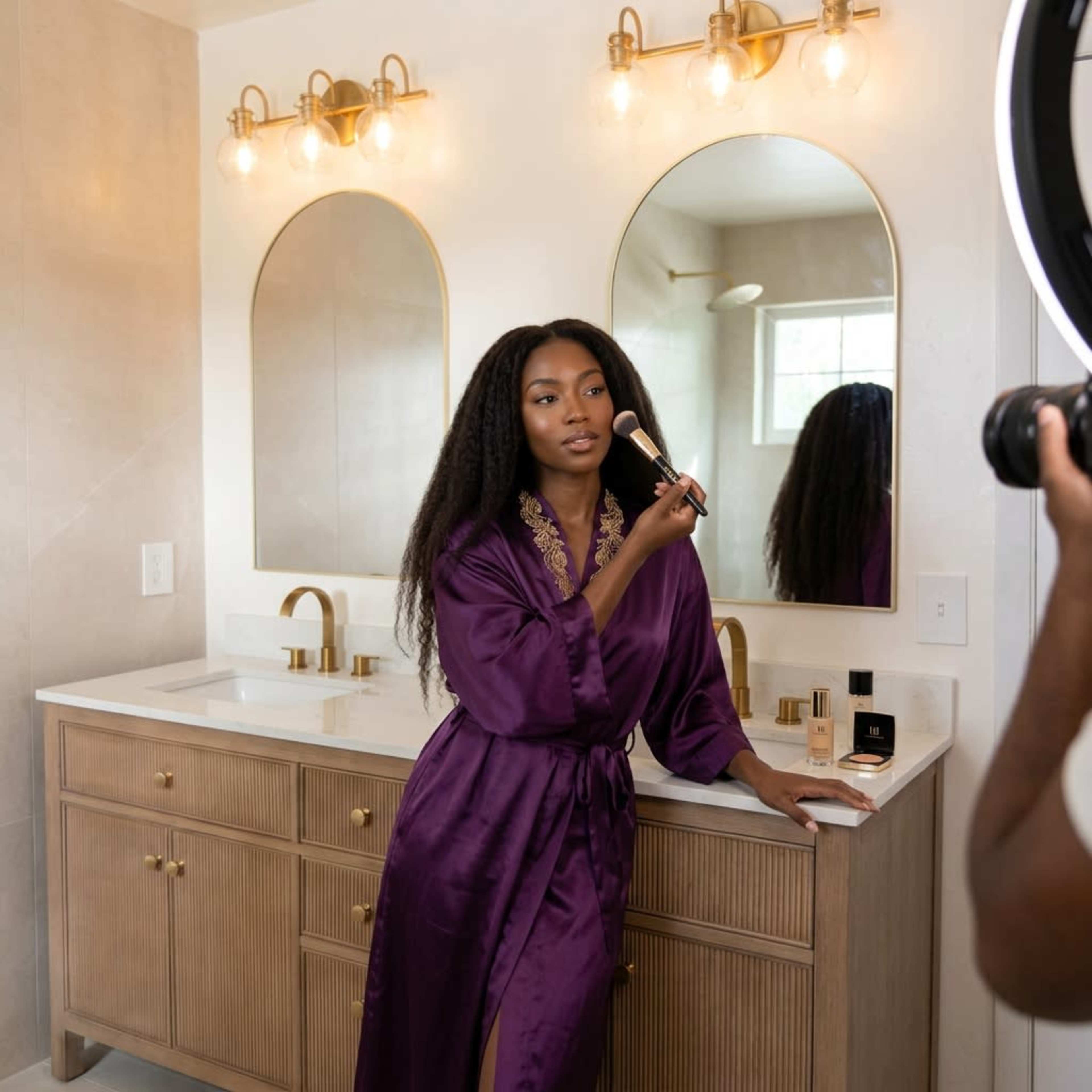 A person in a purple robe is applying makeup in front of a bathroom mirror with stylish lighting.