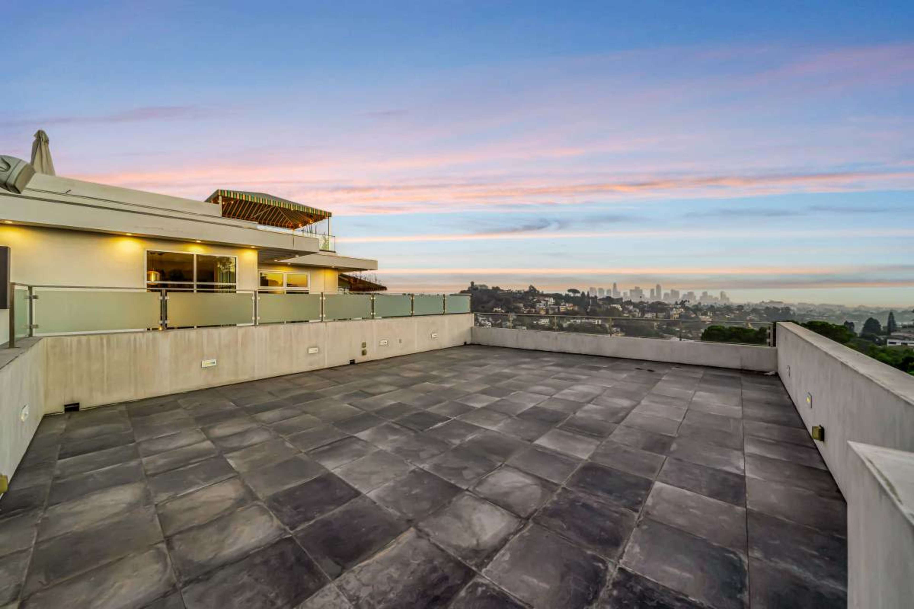 The image shows a spacious rooftop terrace with a tile floor, offering a view of a city skyline against a colorful sunset.