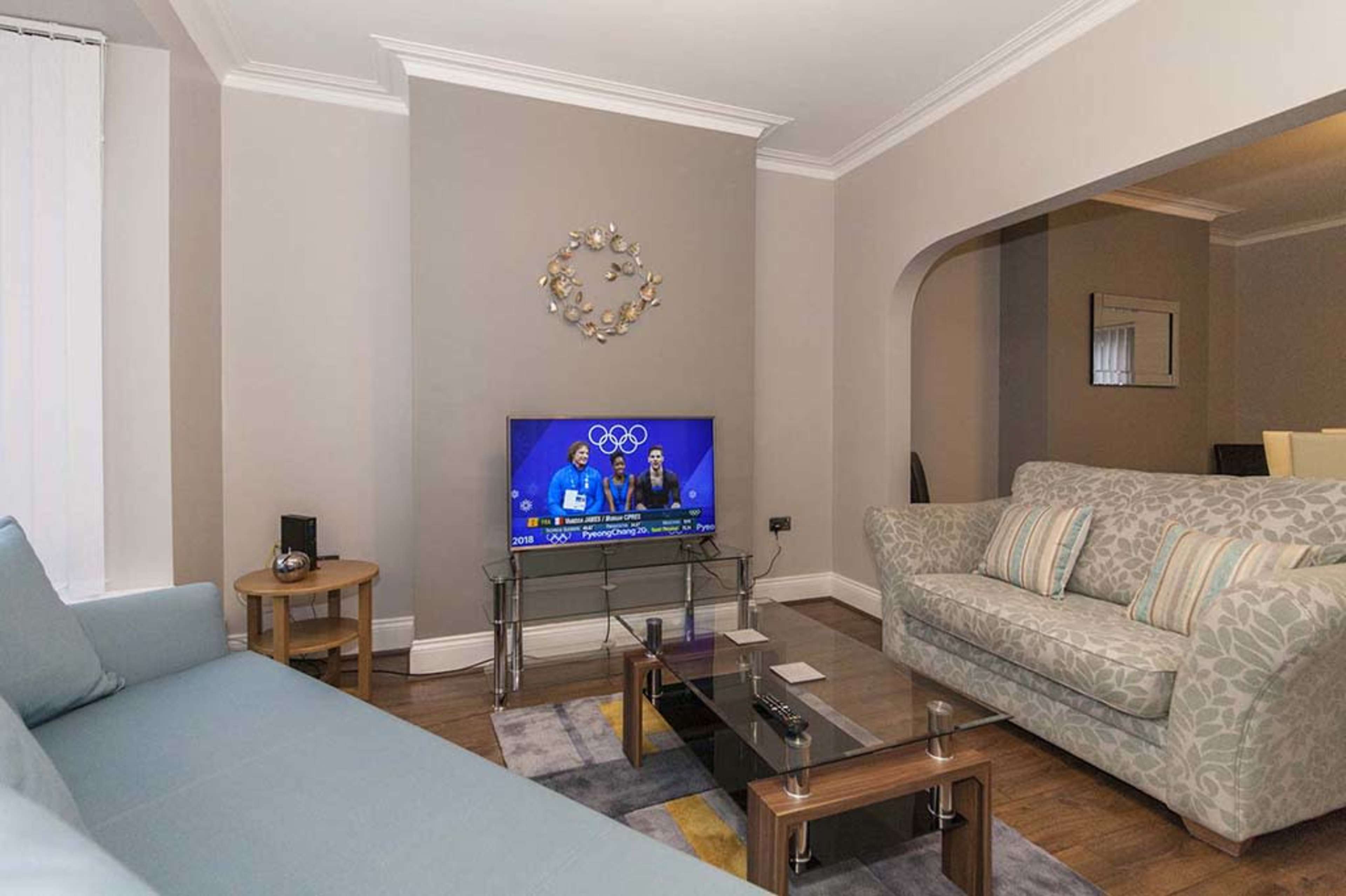 A living room with a light-colored sofa, a patterned couch, a glass coffee table, and a television displaying an Olympic event.