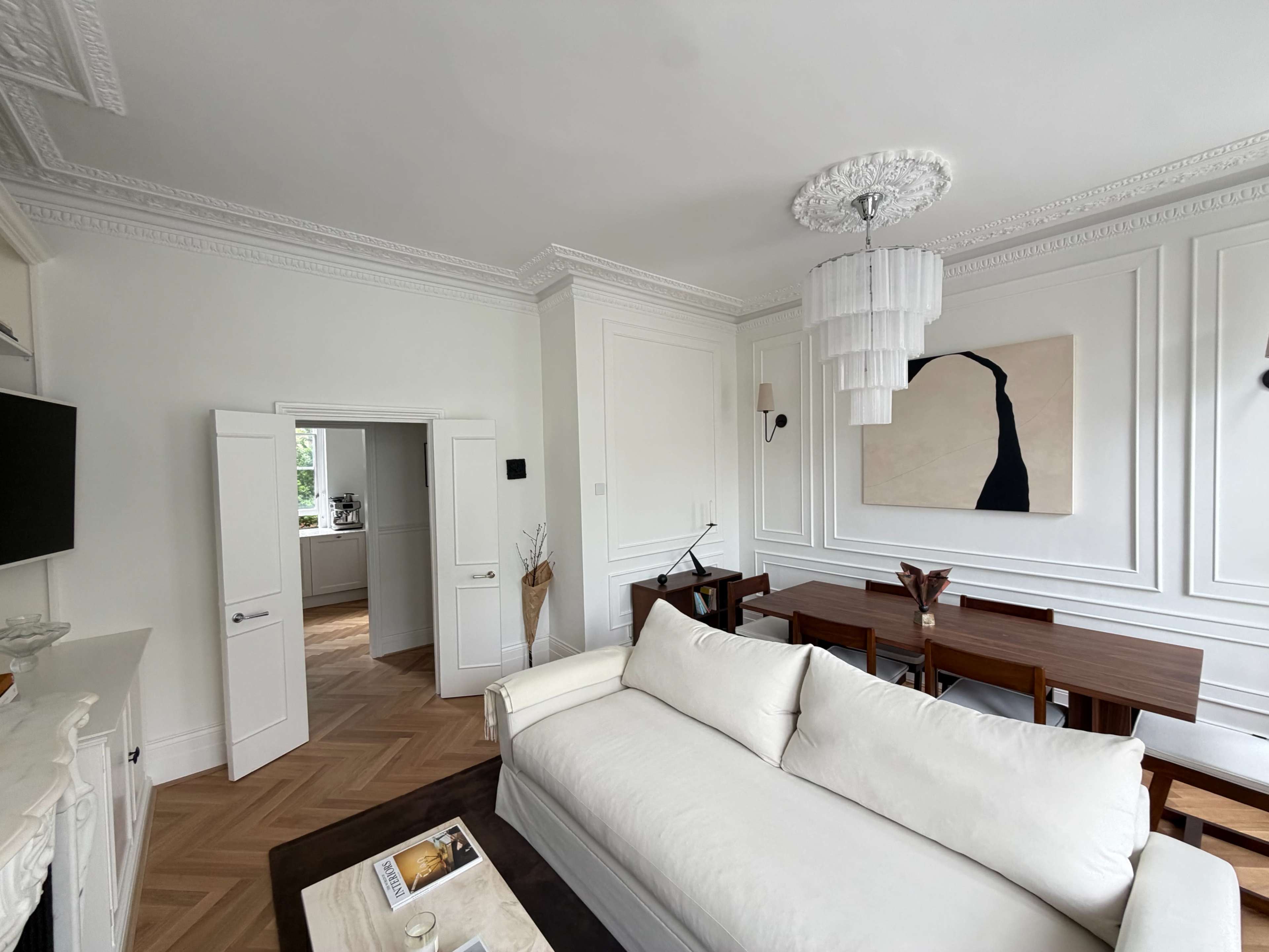 A modern living room features a white sofa, a dining area with wooden furniture, and a large wall art piece.