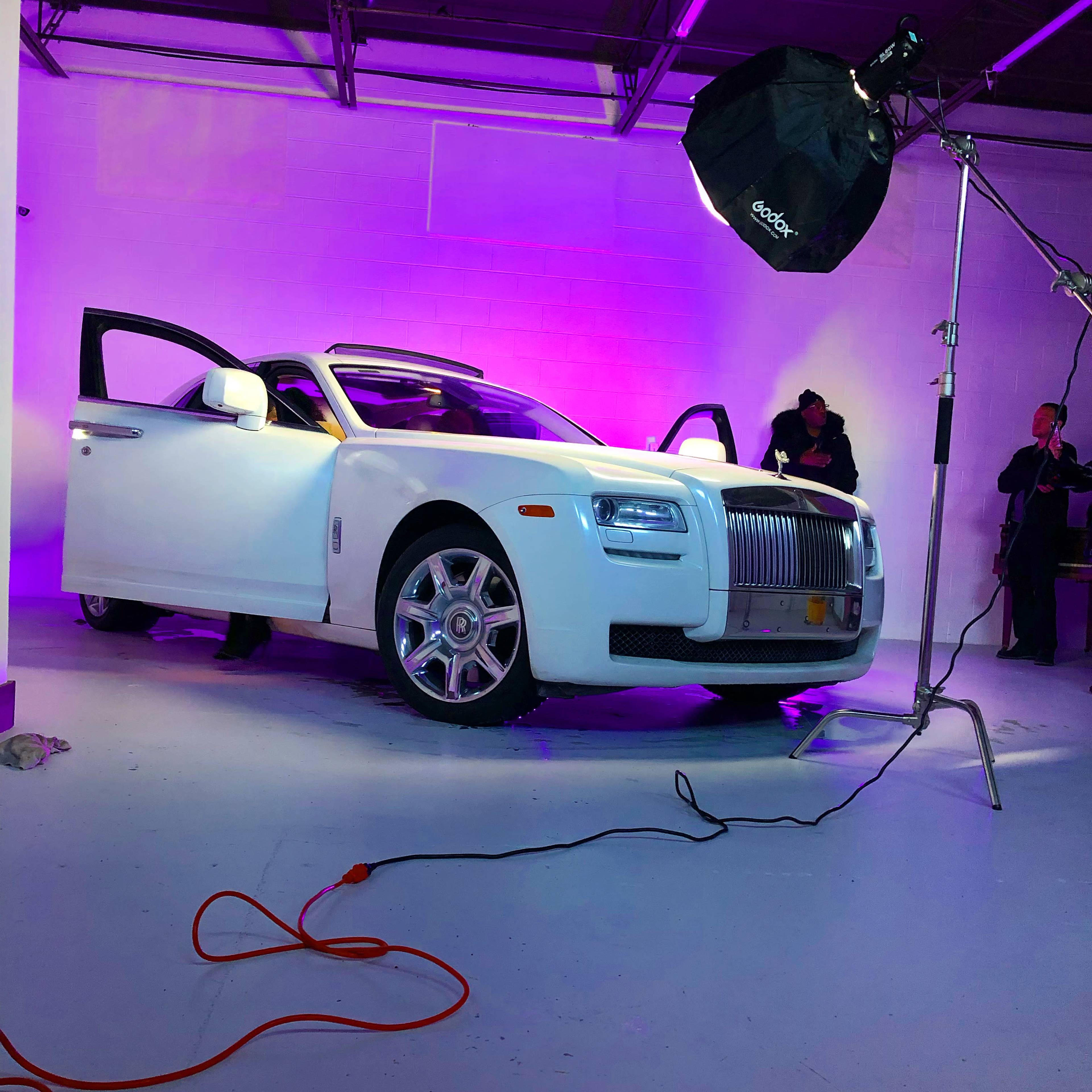 A white luxury car is positioned in a studio with purple lighting, while a photographer adjusts equipment nearby.