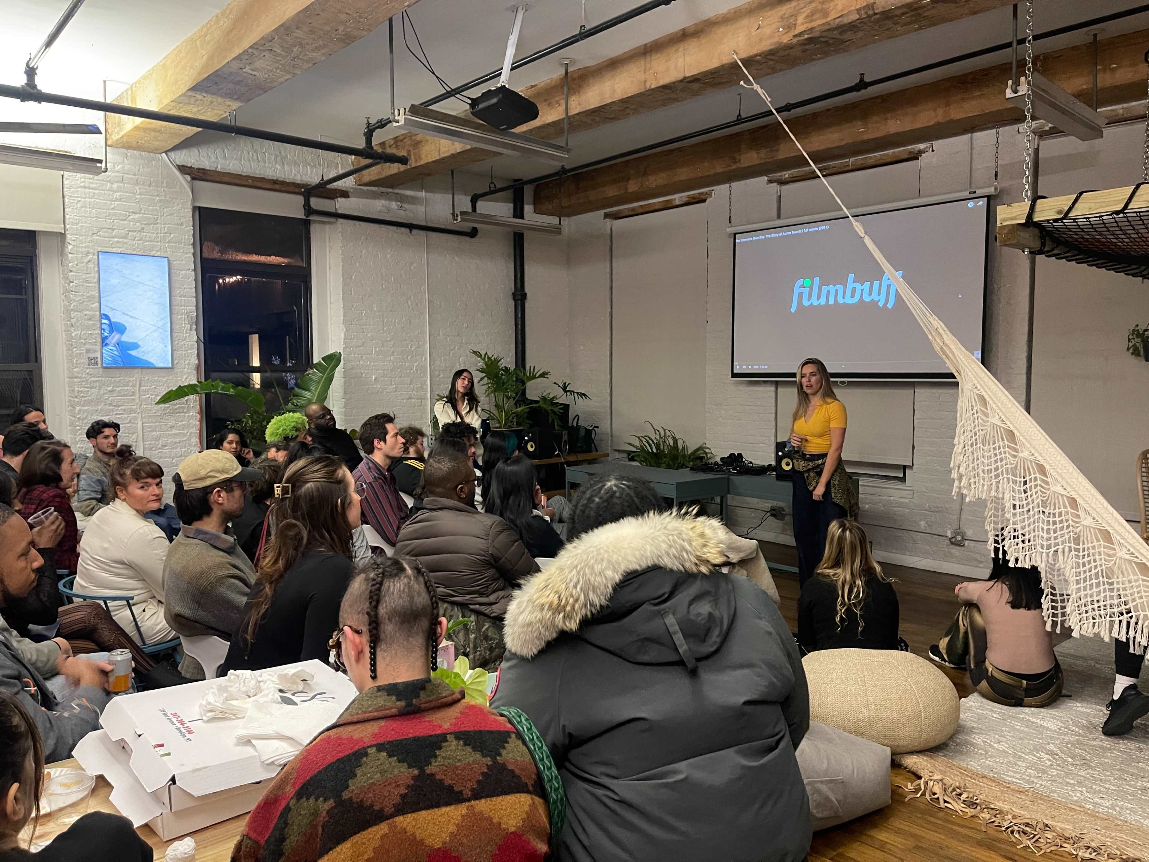 A group of people sits in a modern, well-lit space, attentively watching a presentation on a screen and surrounded by plants and casual seating.