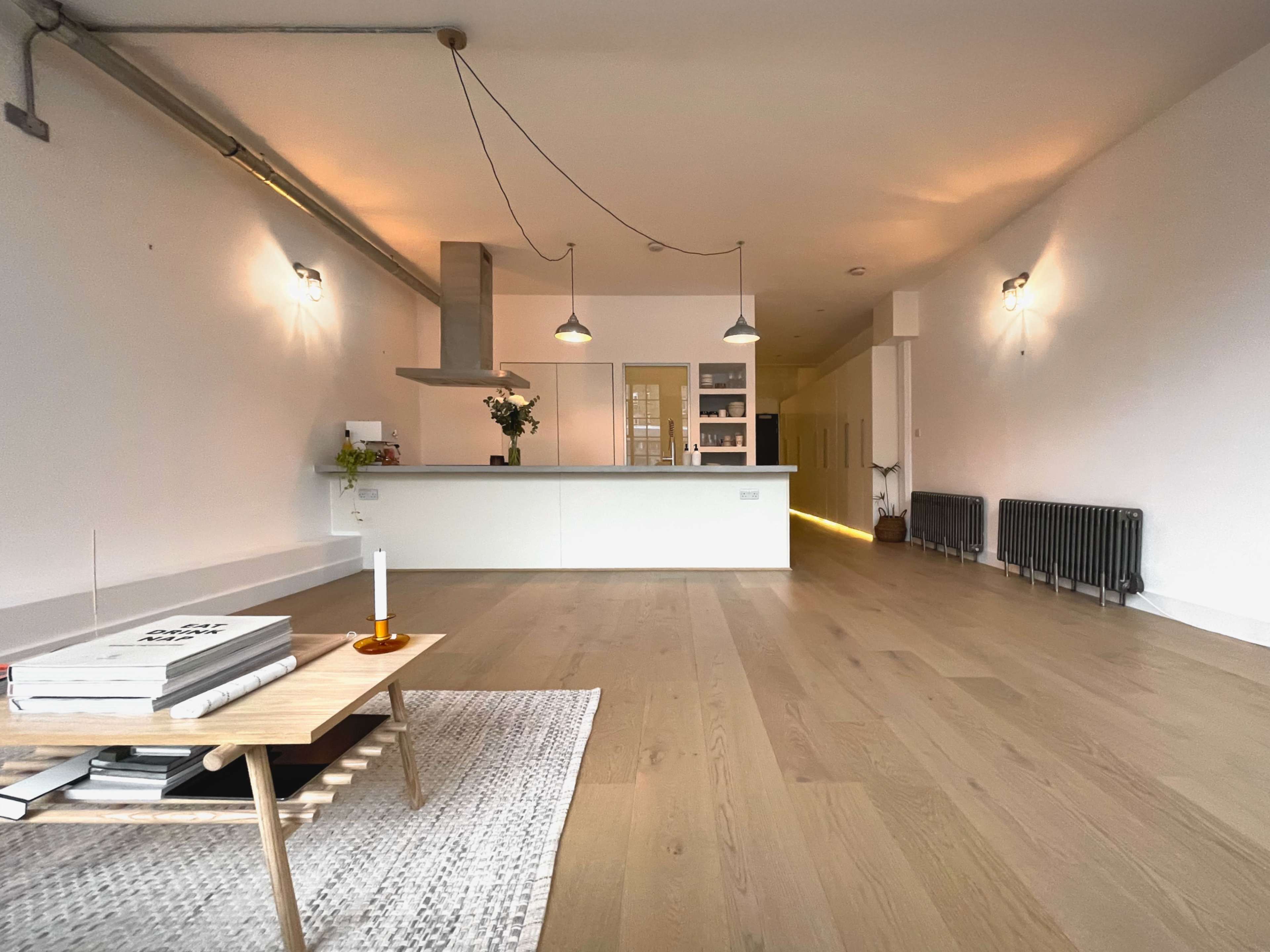 A spacious modern interior features a minimalist kitchen and living area with wooden flooring, a coffee table, and radiant lighting.