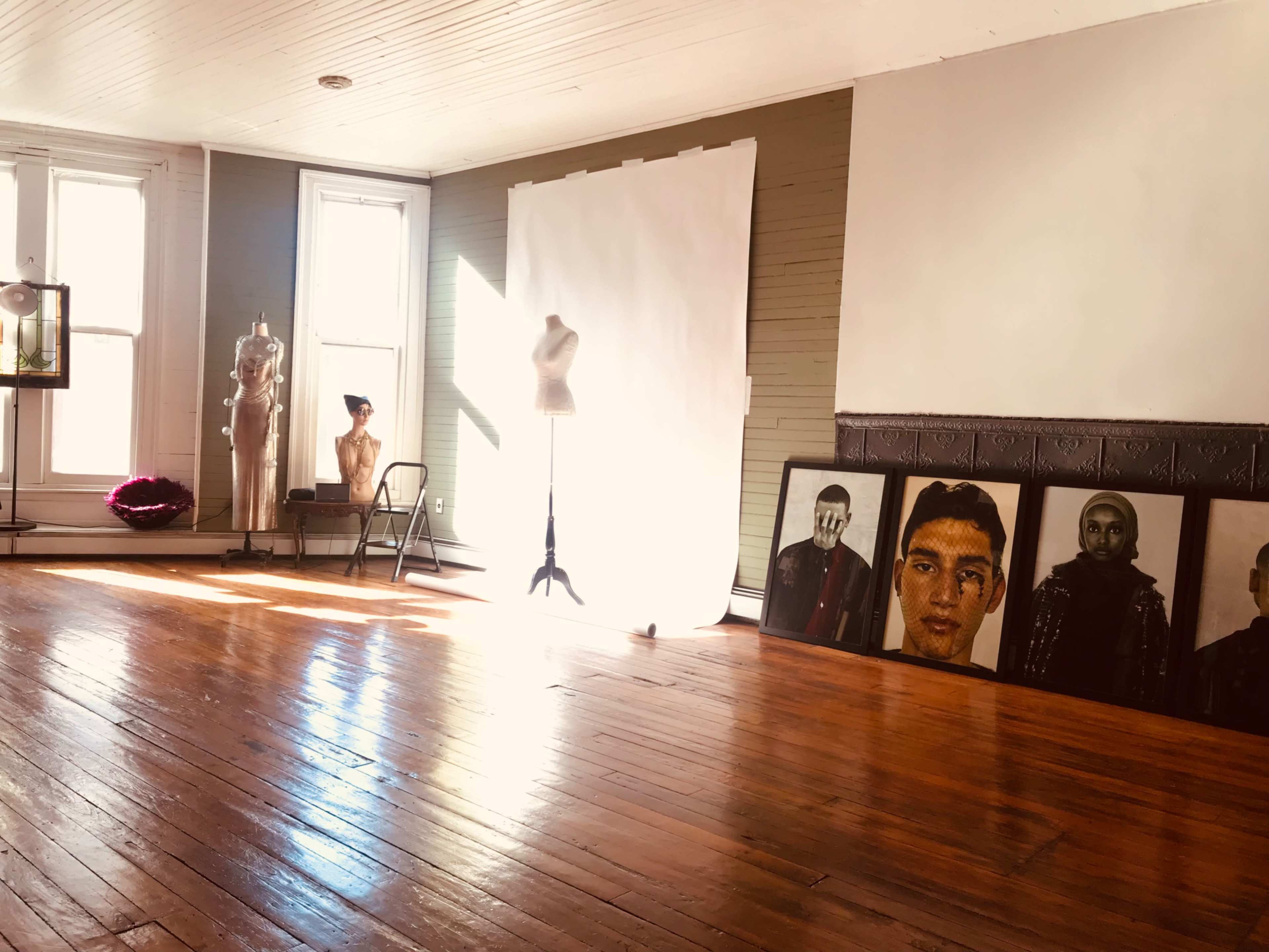 A spacious artist studio with wooden floors, large windows, and a backdrop alongside mannequins and portraits displayed against the wall.