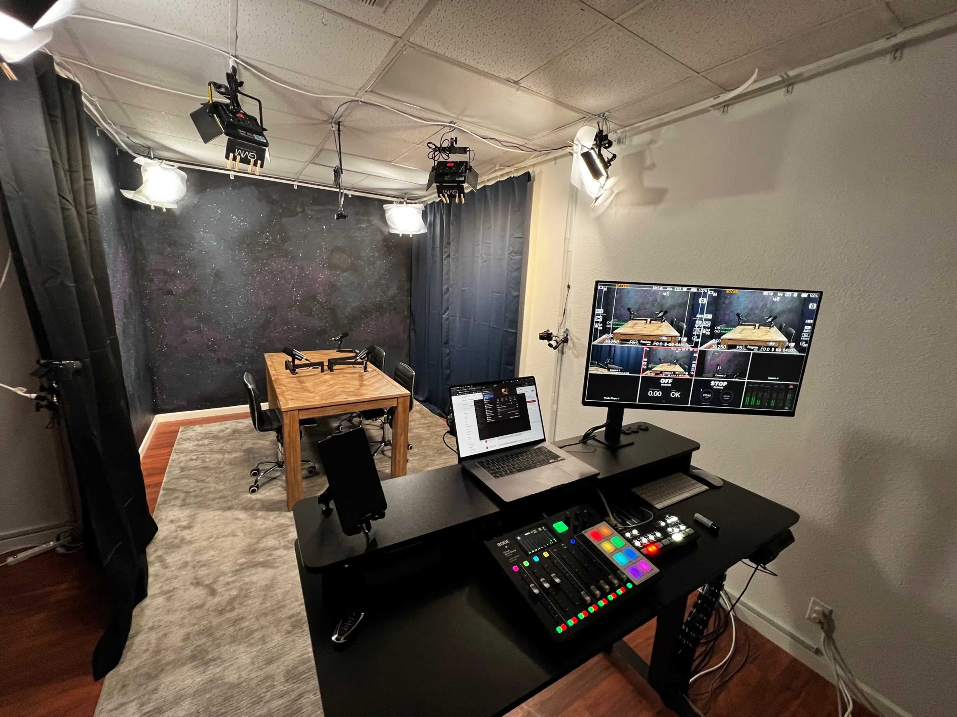 A video production studio features a control desk with a laptop and monitoring equipment, surrounded by a table and chairs, illuminated by studio lights.