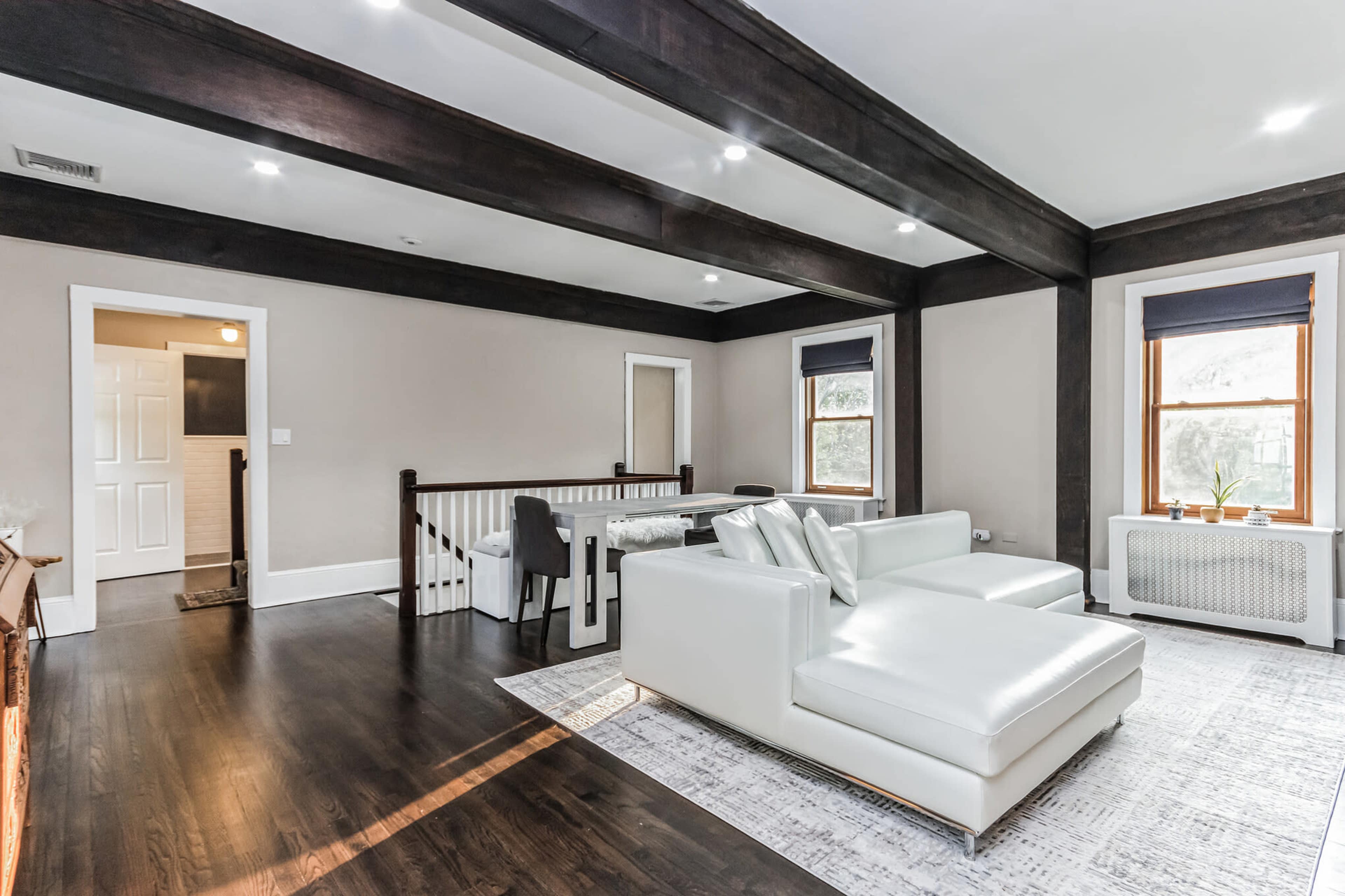 The image shows a spacious living room with dark wooden beams, a white sectional sofa, and large windows featuring light-colored shades.