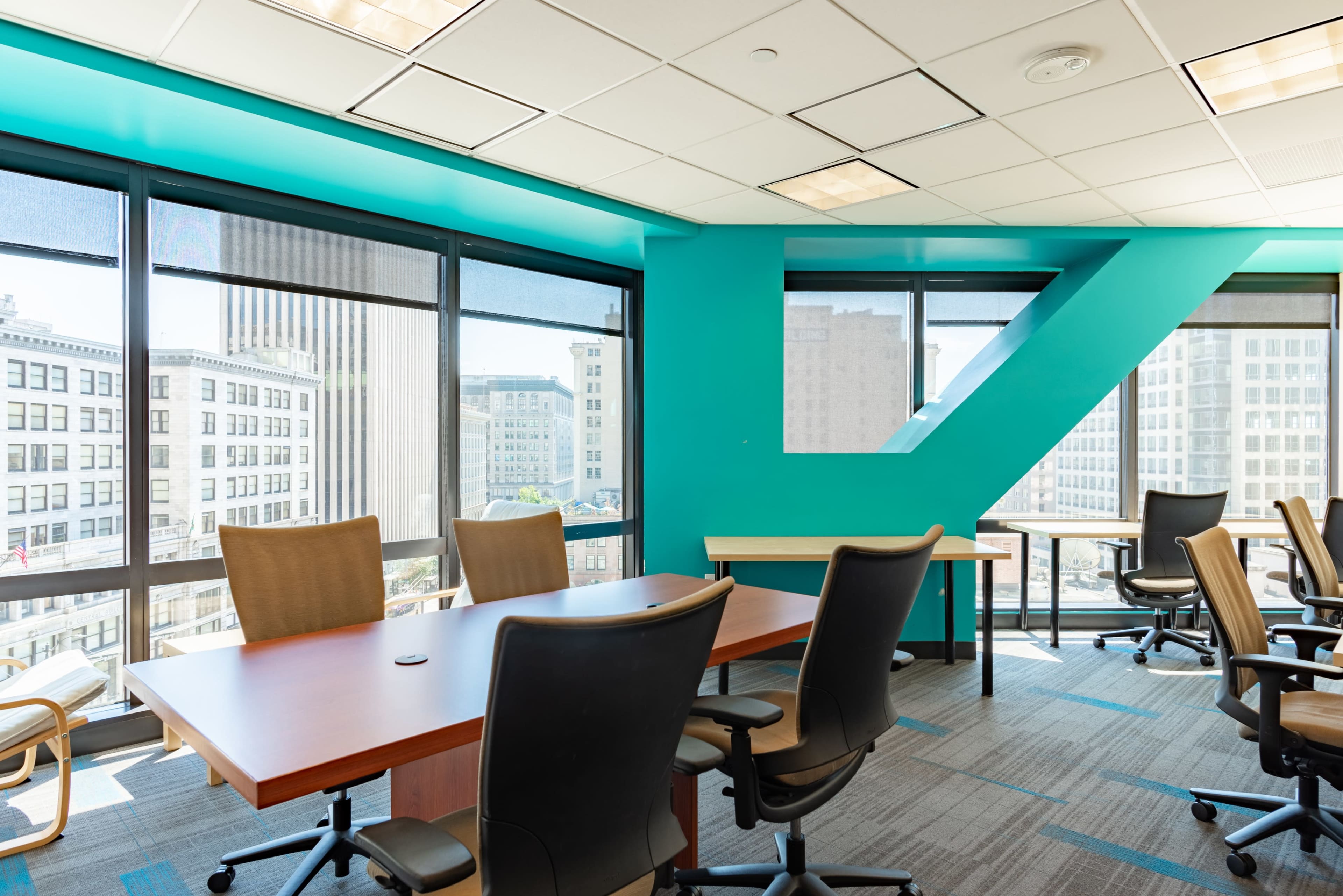 A modern conference room features a large wooden table surrounded by office chairs and large windows with a view of a cityscape.
