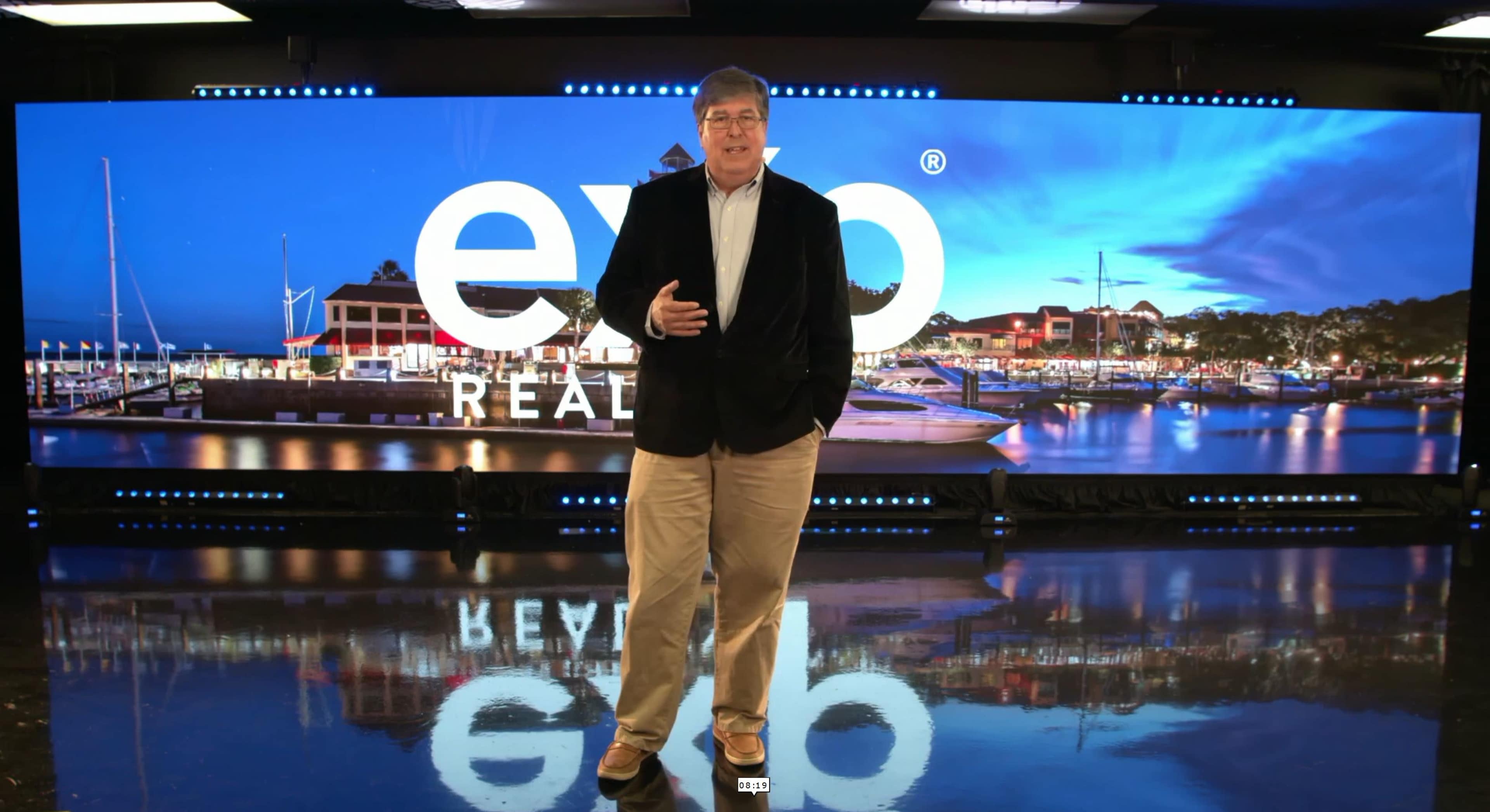 A man stands in front of a digital backdrop featuring a marina with boats and buildings, promoting a real estate brand.
