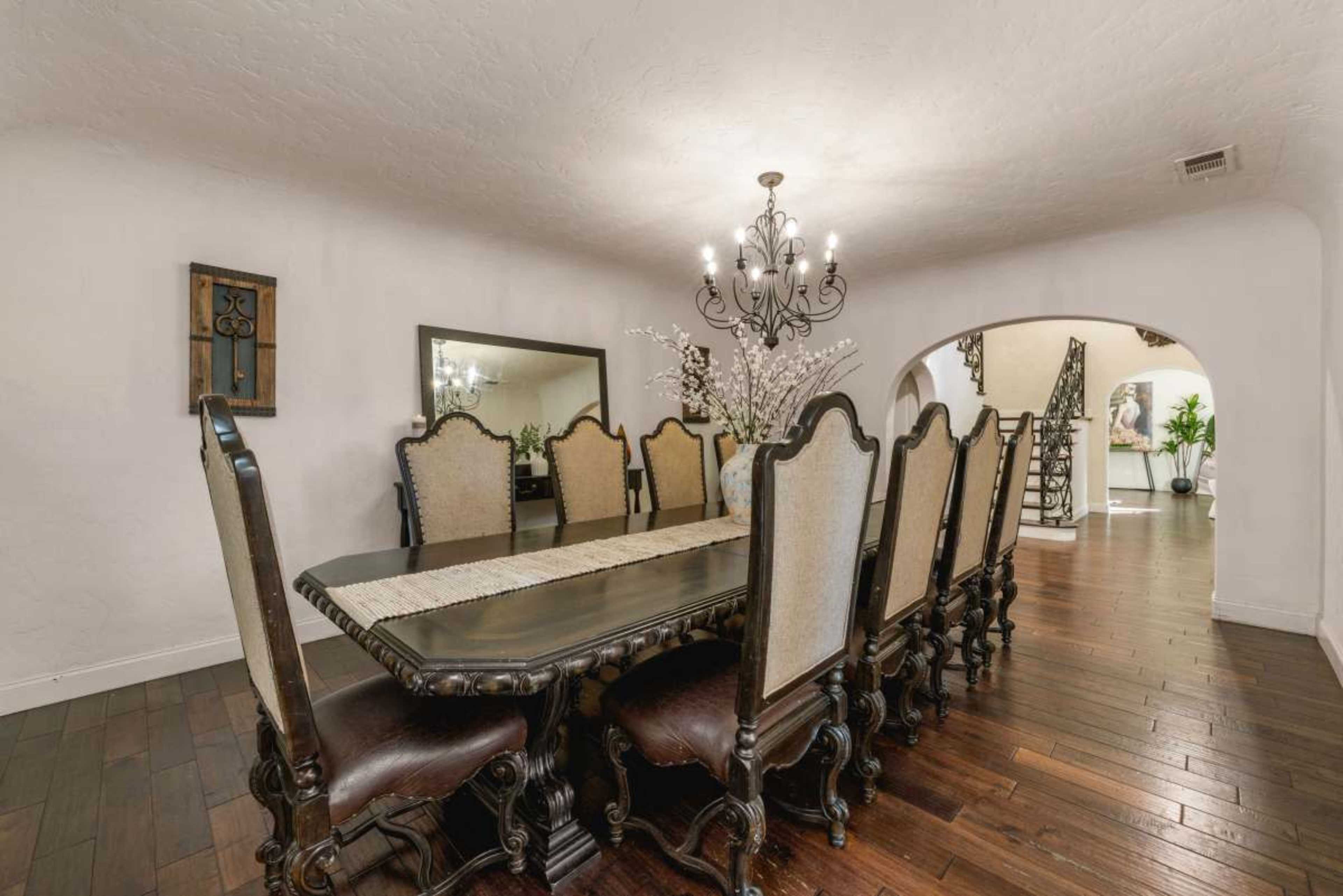 The image shows a spacious dining room with a large wooden table surrounded by ten upholstered chairs, accented by a chandelier and a decorative mirror on the wall.
