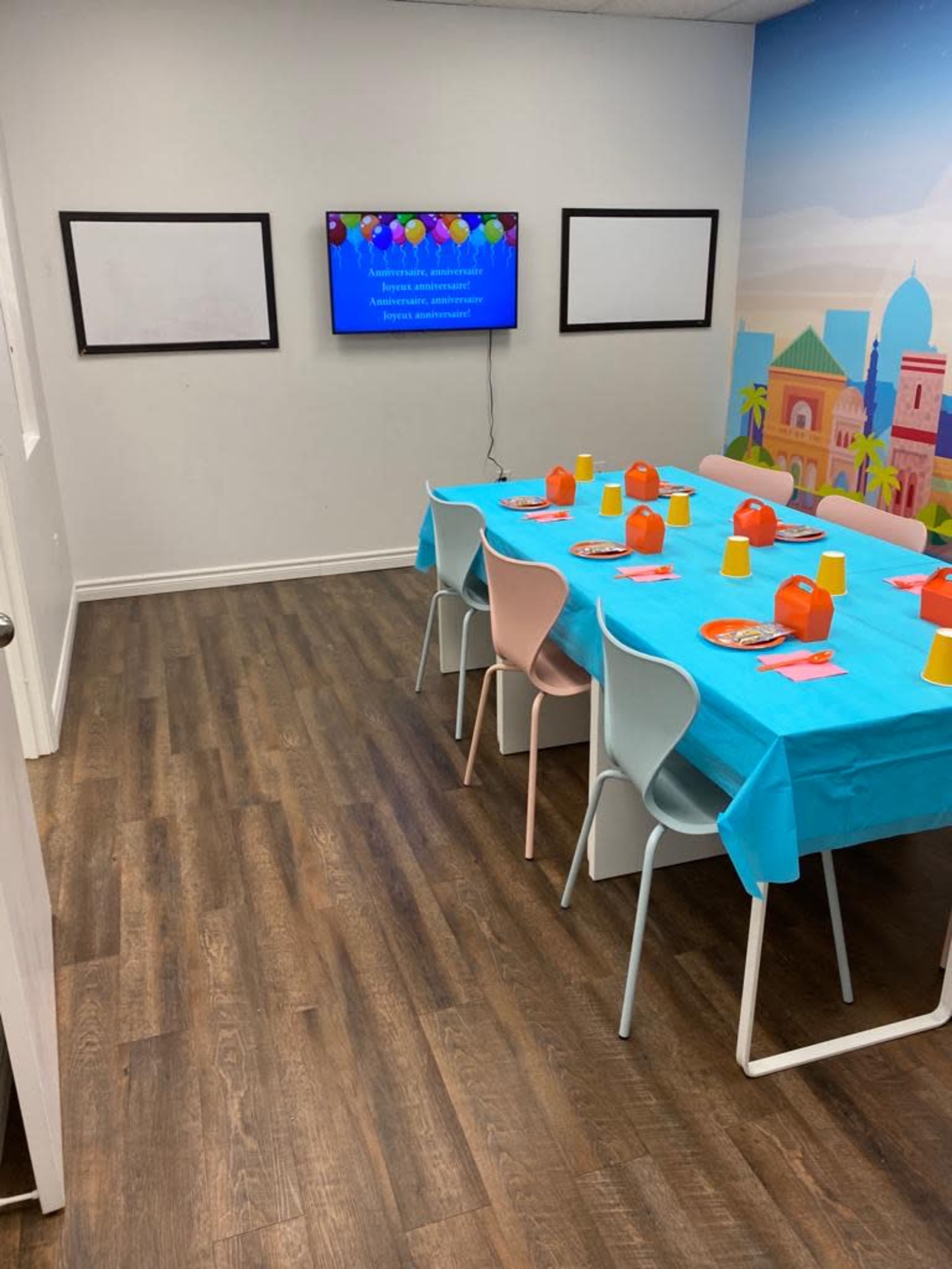 A brightly colored party room features a table set with plates and cups, with a television displaying decorations on one wall.
