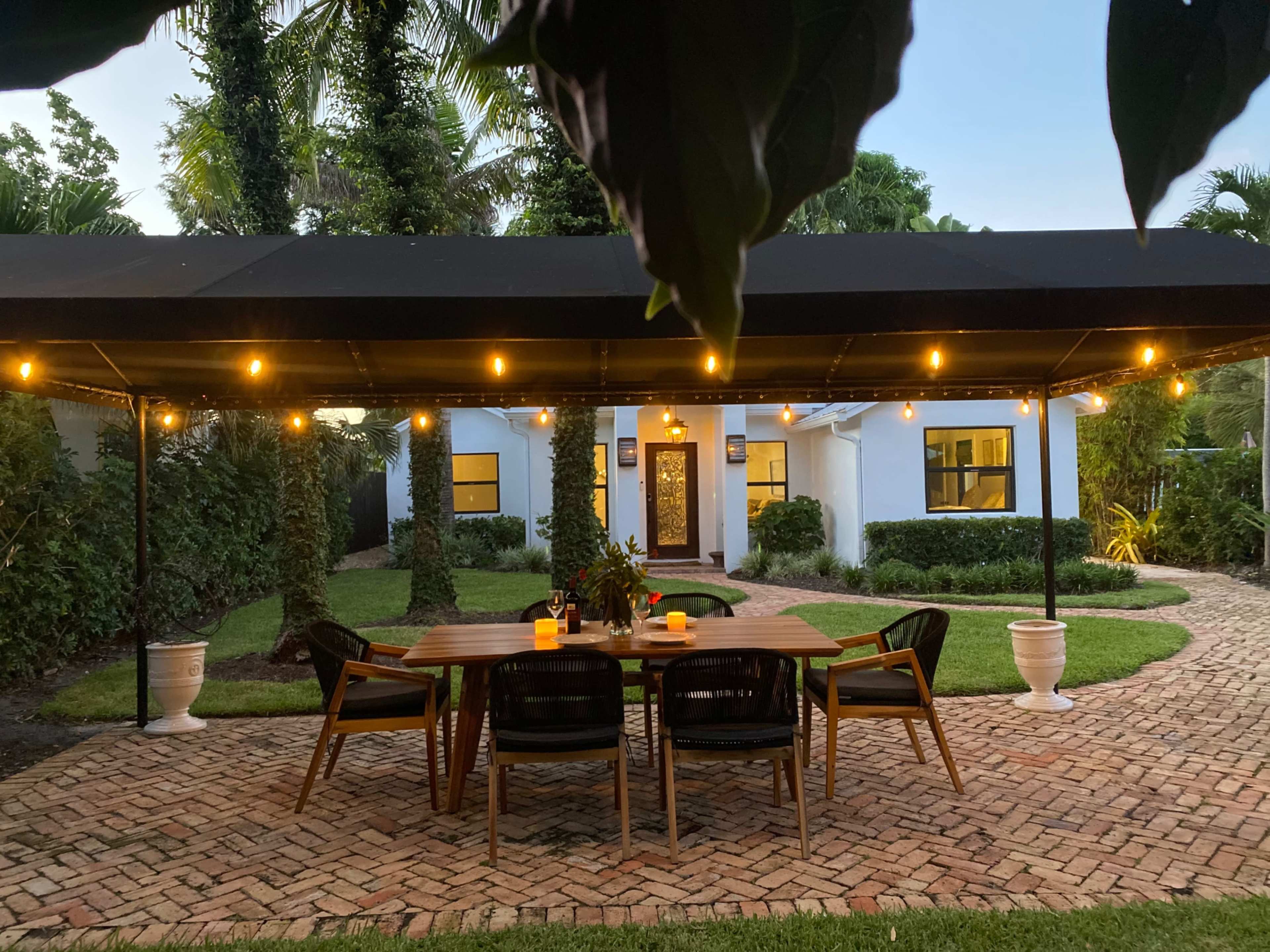 A patio with a wooden table and chairs is illuminated by string lights, leading to a house surrounded by greenery.