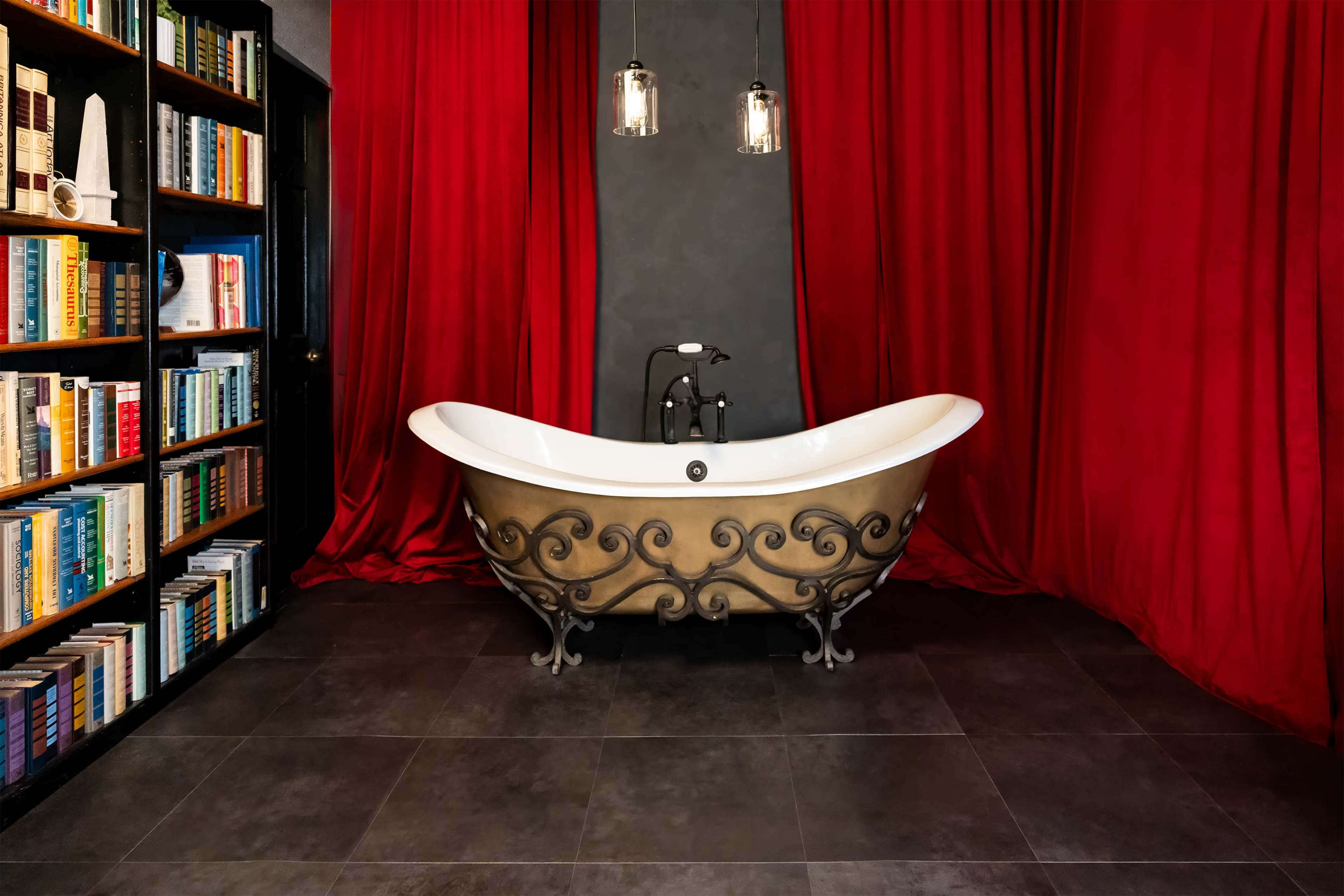 A vintage clawfoot bathtub is positioned in front of a dark wall, surrounded by red curtains and flanked by a bookshelf filled with books.
