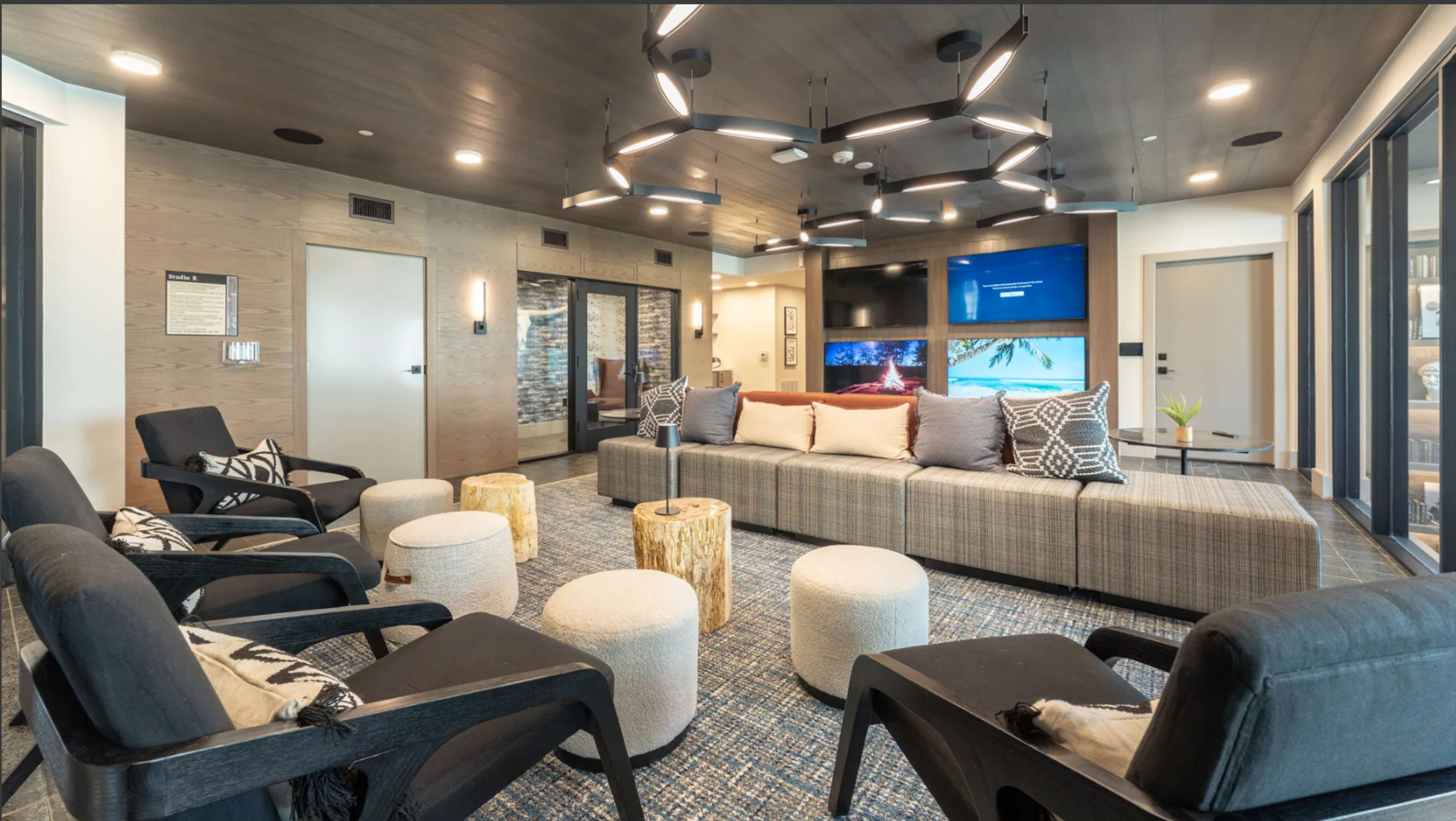 The image shows a modern lounge area featuring a sectional sofa, circular stools, and dark chairs under artistic lighting, with a large screen displaying a scenic image on one wall.