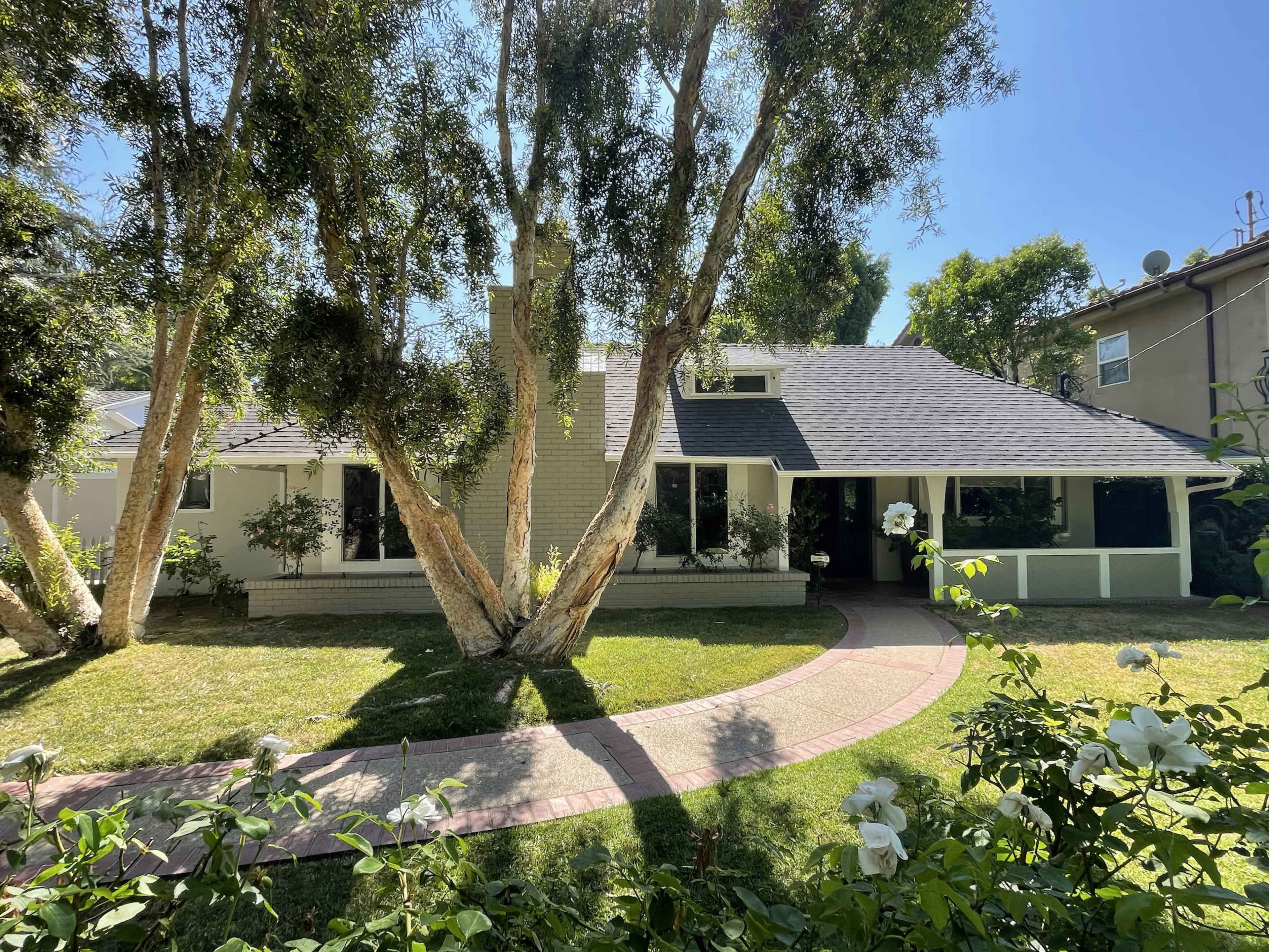 A single-story house with a dark roof and large windows is set in a green yard with trees and a curved walkway.
