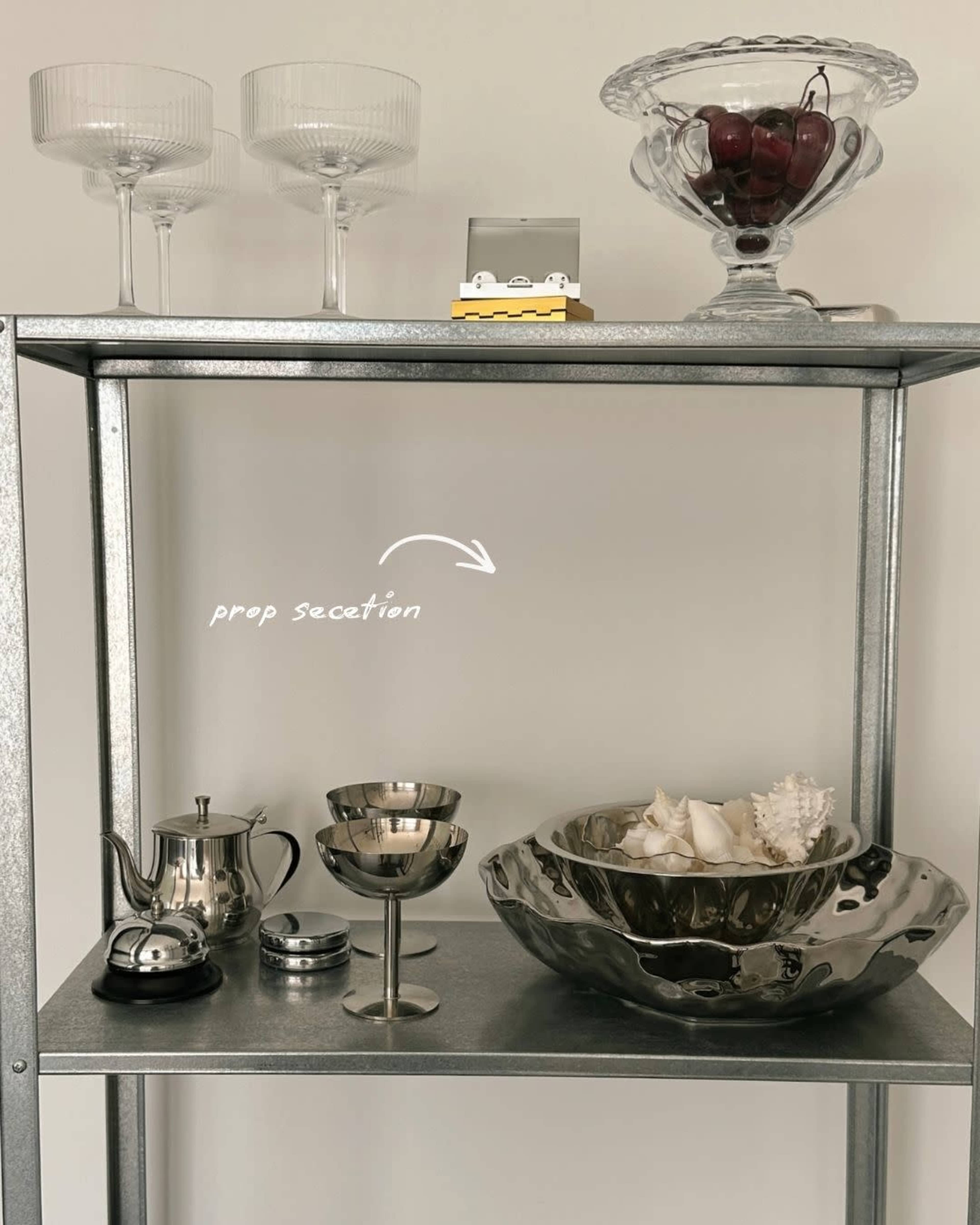 A metallic shelf displays various kitchen items, including a teapot, wine glasses, a bowl of shells, and a glass dish filled with grapes.