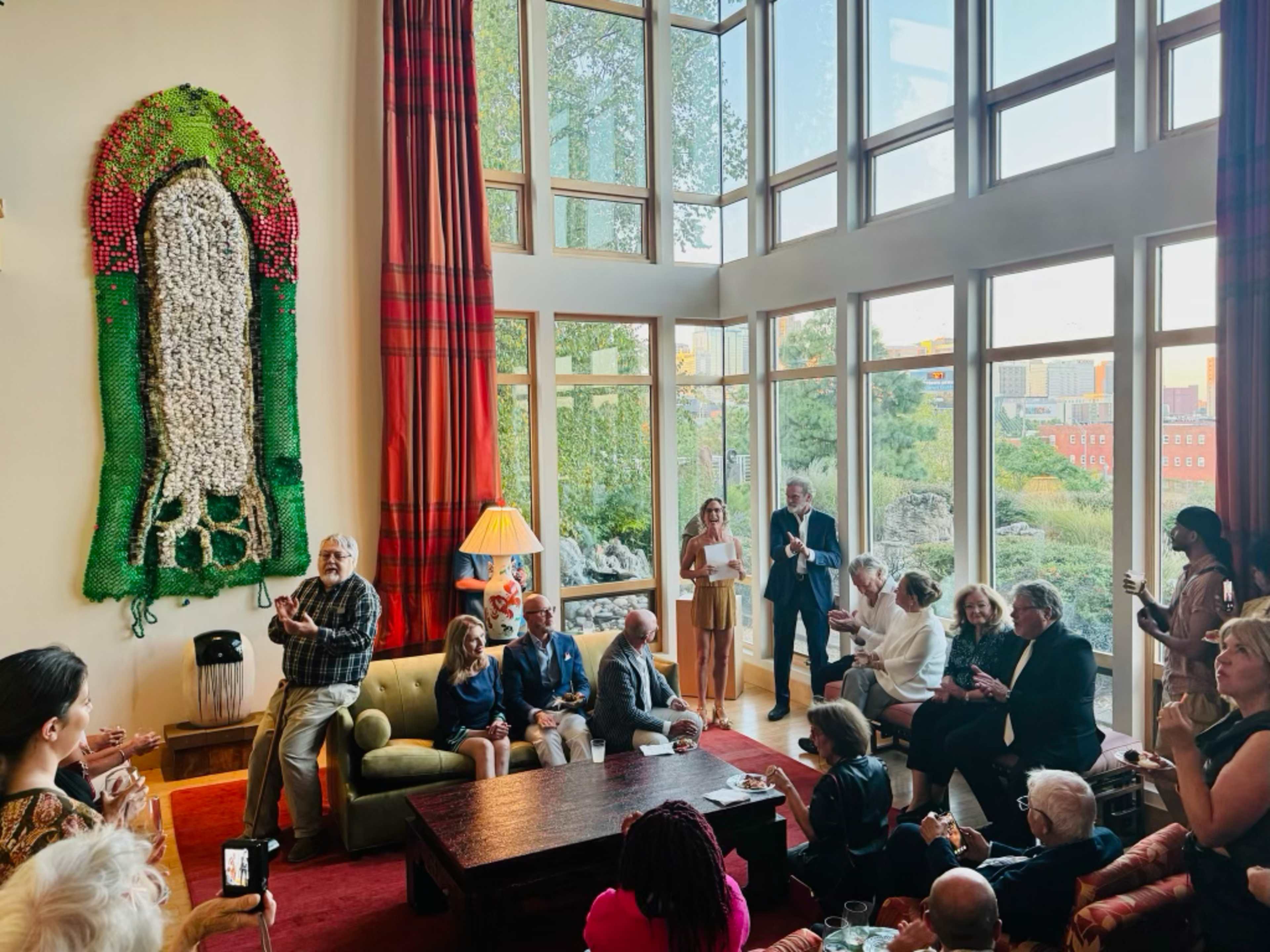 A group of people are seated and standing in a brightly lit room with large windows, surrounded by greenery and a decorative wall piece made of various materials.