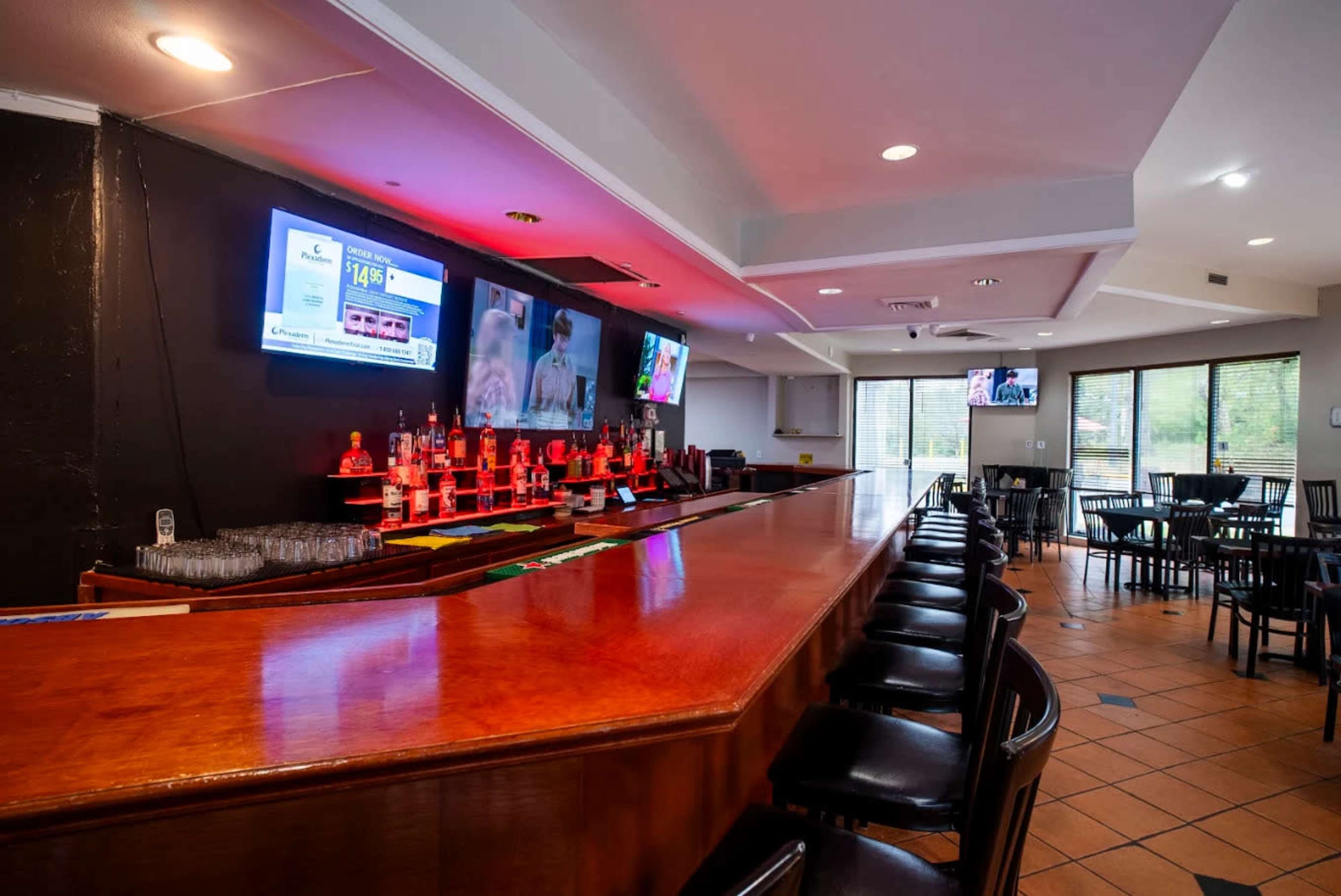 The image shows a well-lit bar area with a polished wooden counter, multiple bottles arranged on shelves, and two large screens displaying content on the wall.