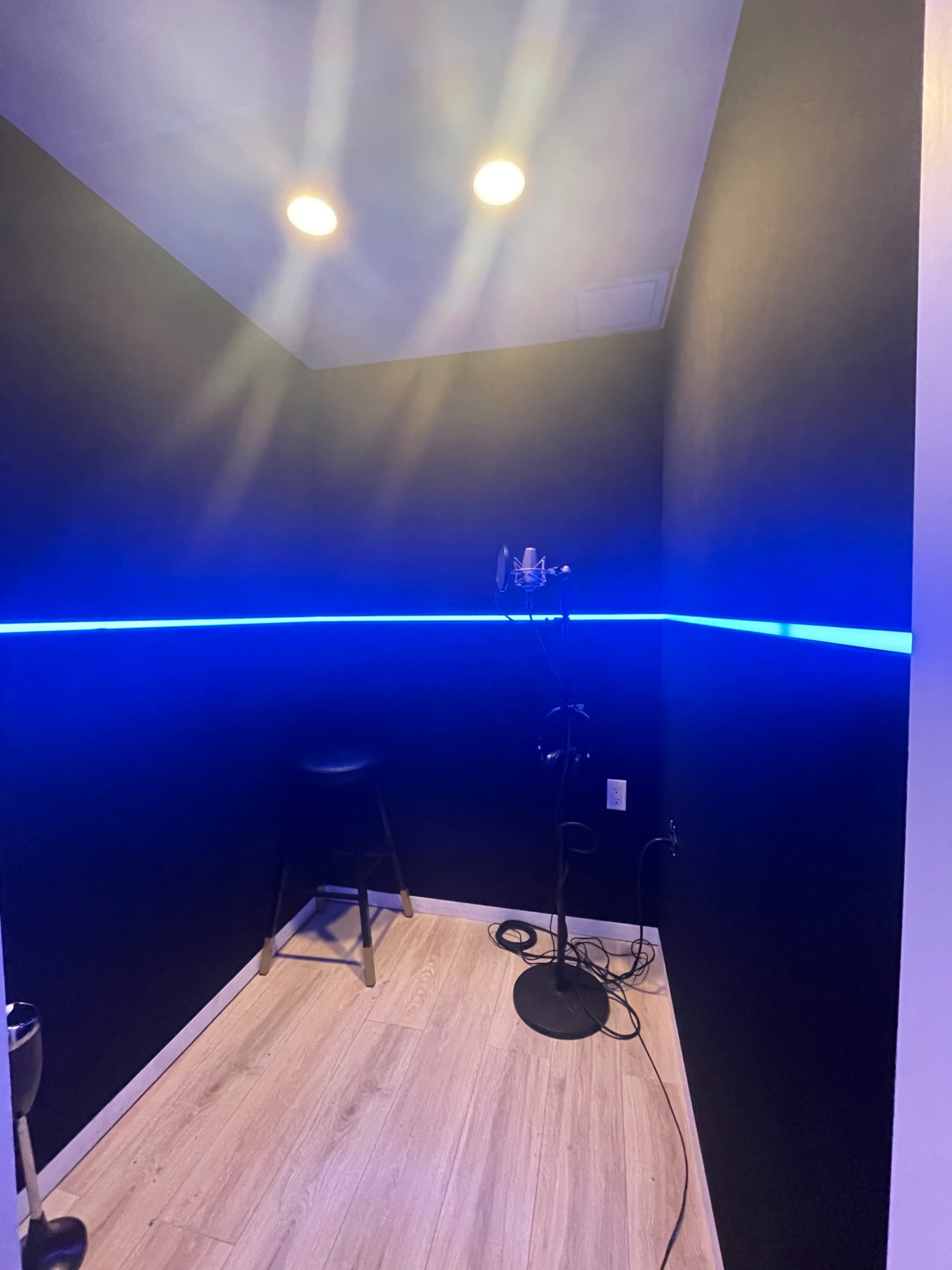 A small recording booth featuring black walls, blue LED lighting, a microphone on a stand, and a stool.