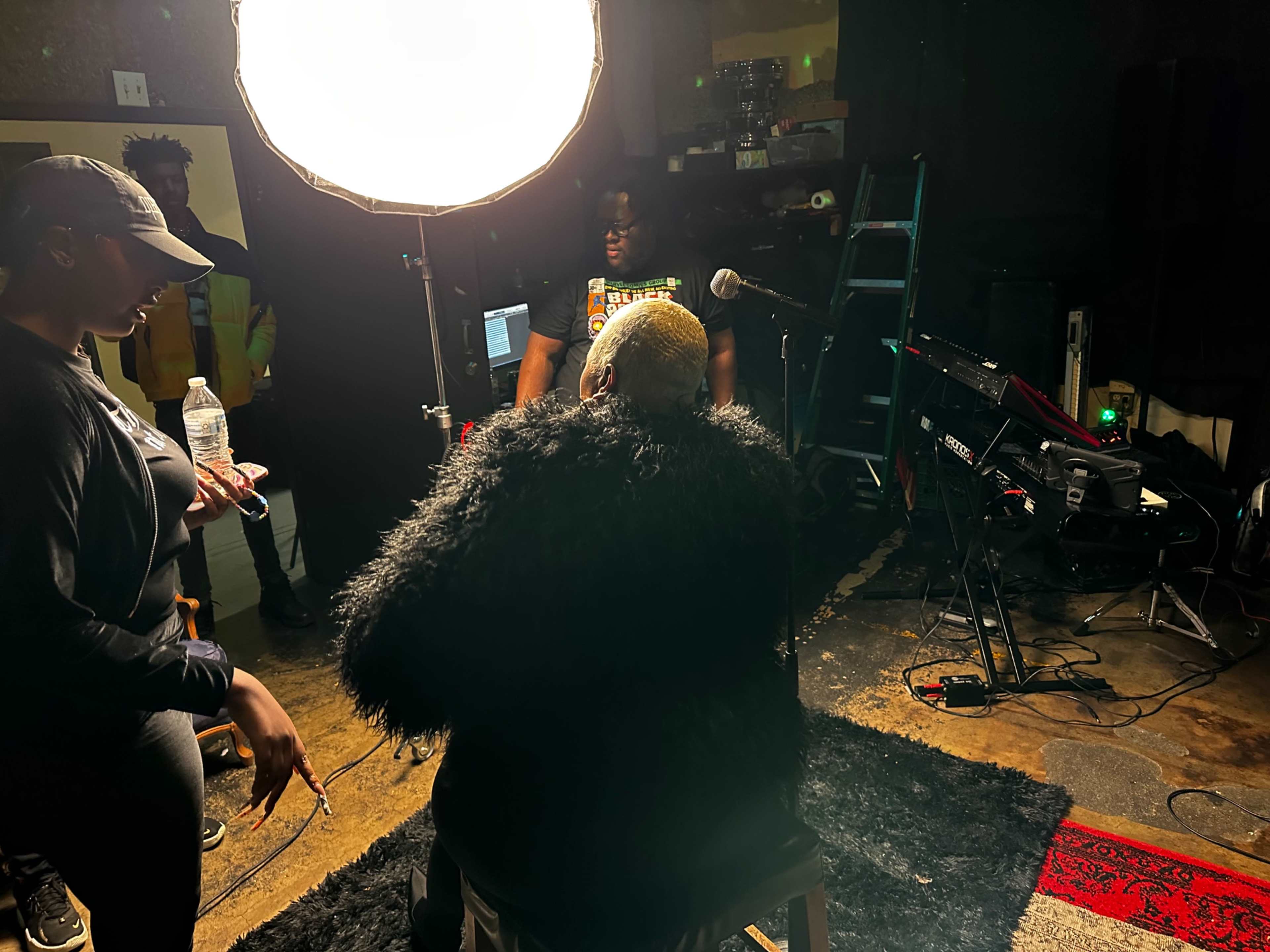 A person wearing a fluffy black garment sits on a stool in a dimly lit room with music equipment and a large spotlight overhead.