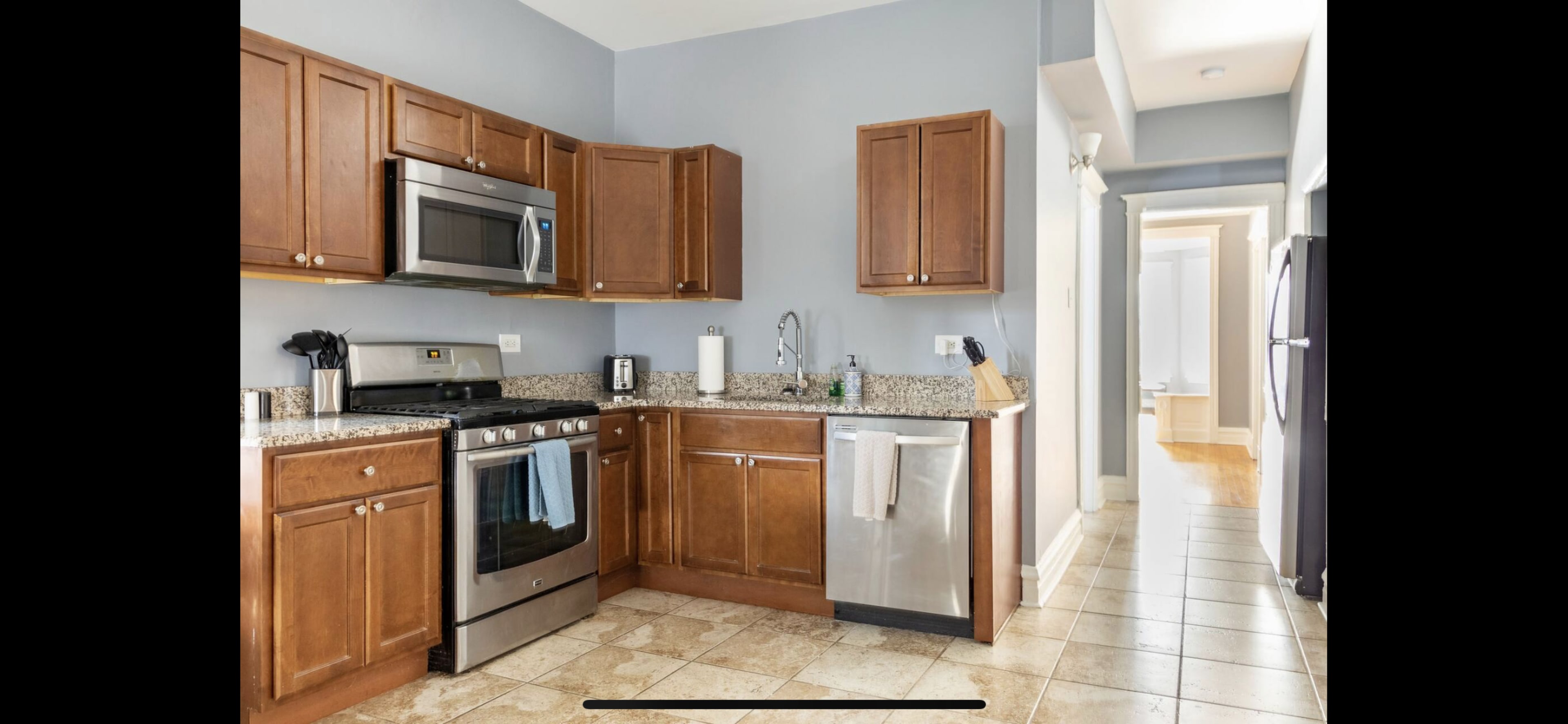 A kitchen with wooden cabinets, stainless steel appliances, and granite countertops, leading to a hallway.