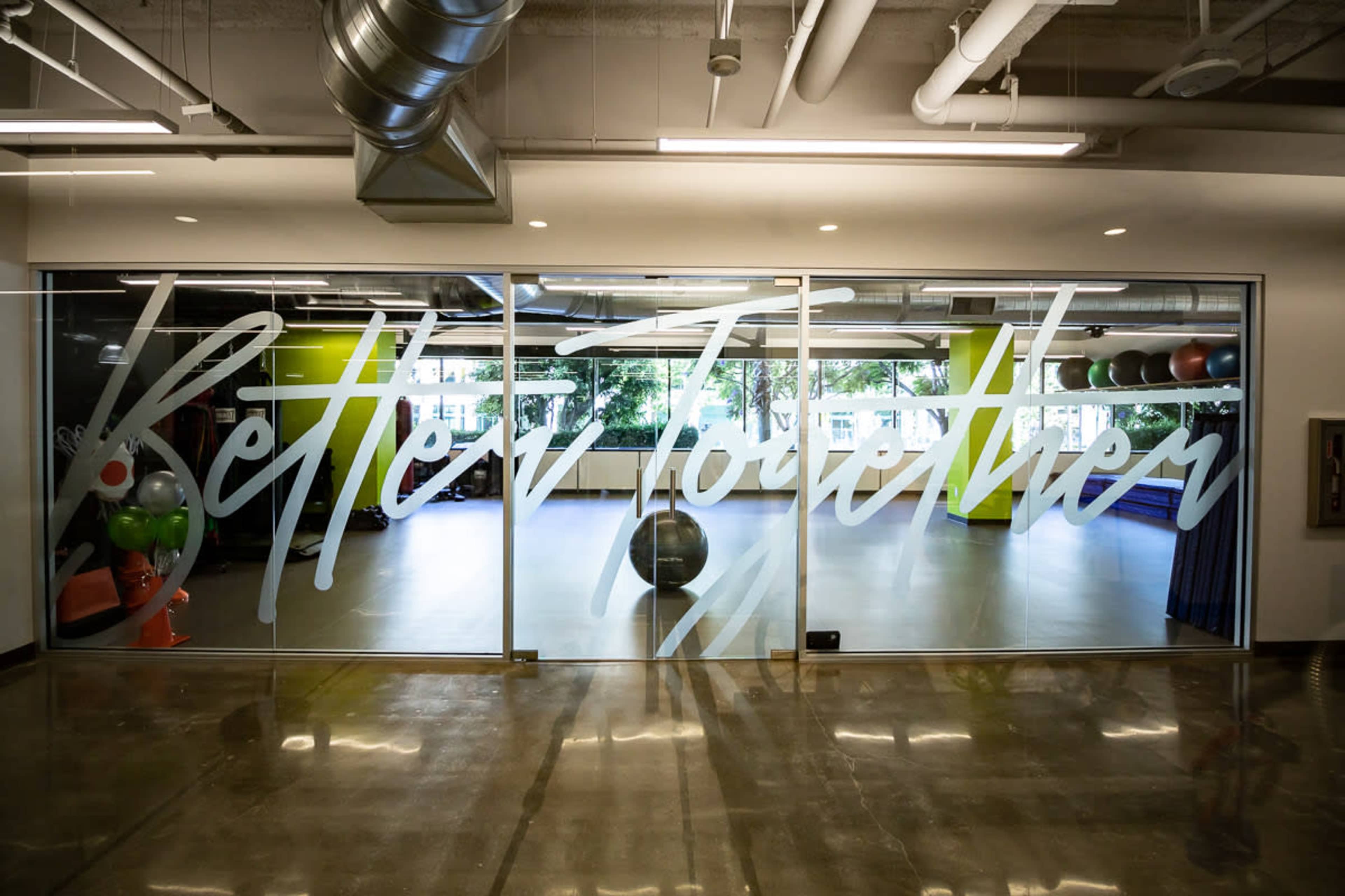 The image shows a glass storefront with the phrase "Better Together" in large, white letters, revealing an interior space equipped with exercise equipment and fitness tools.