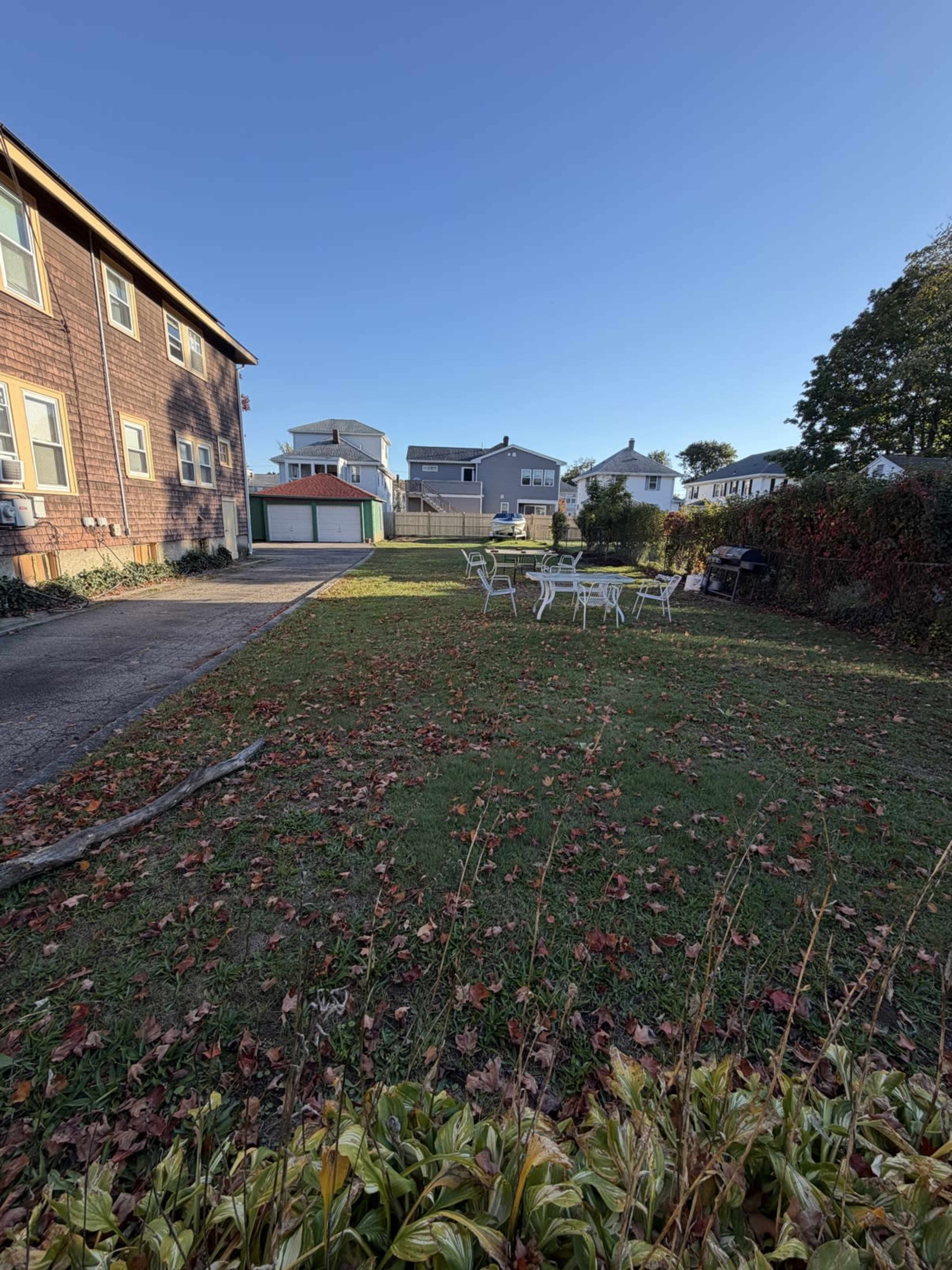 A spacious backyard features a row of white chairs among fallen leaves, with residential buildings and a garage visible in the background.