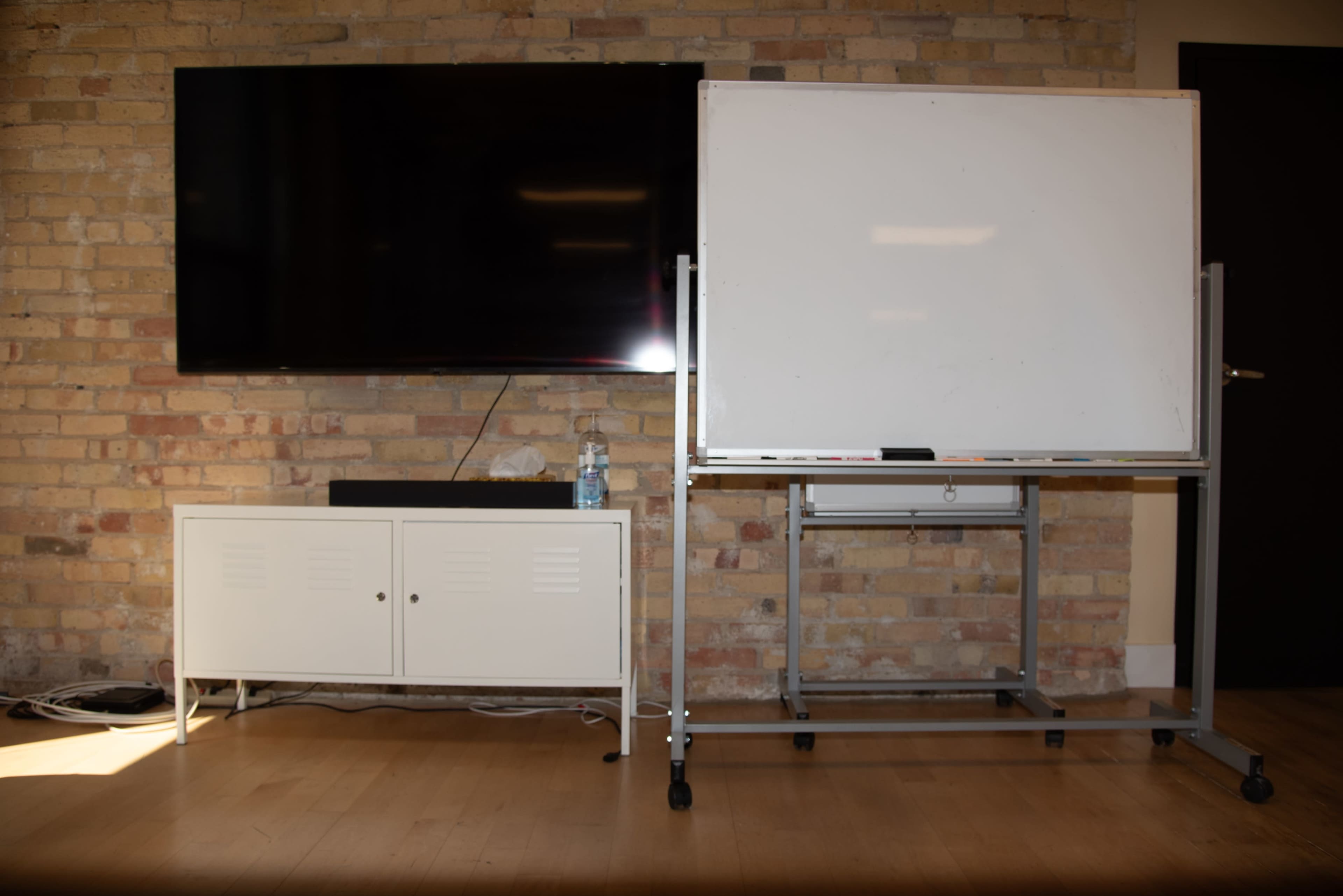 The image shows a television mounted on a brick wall beside a movable whiteboard on a wheeled frame in a room with wooden flooring.