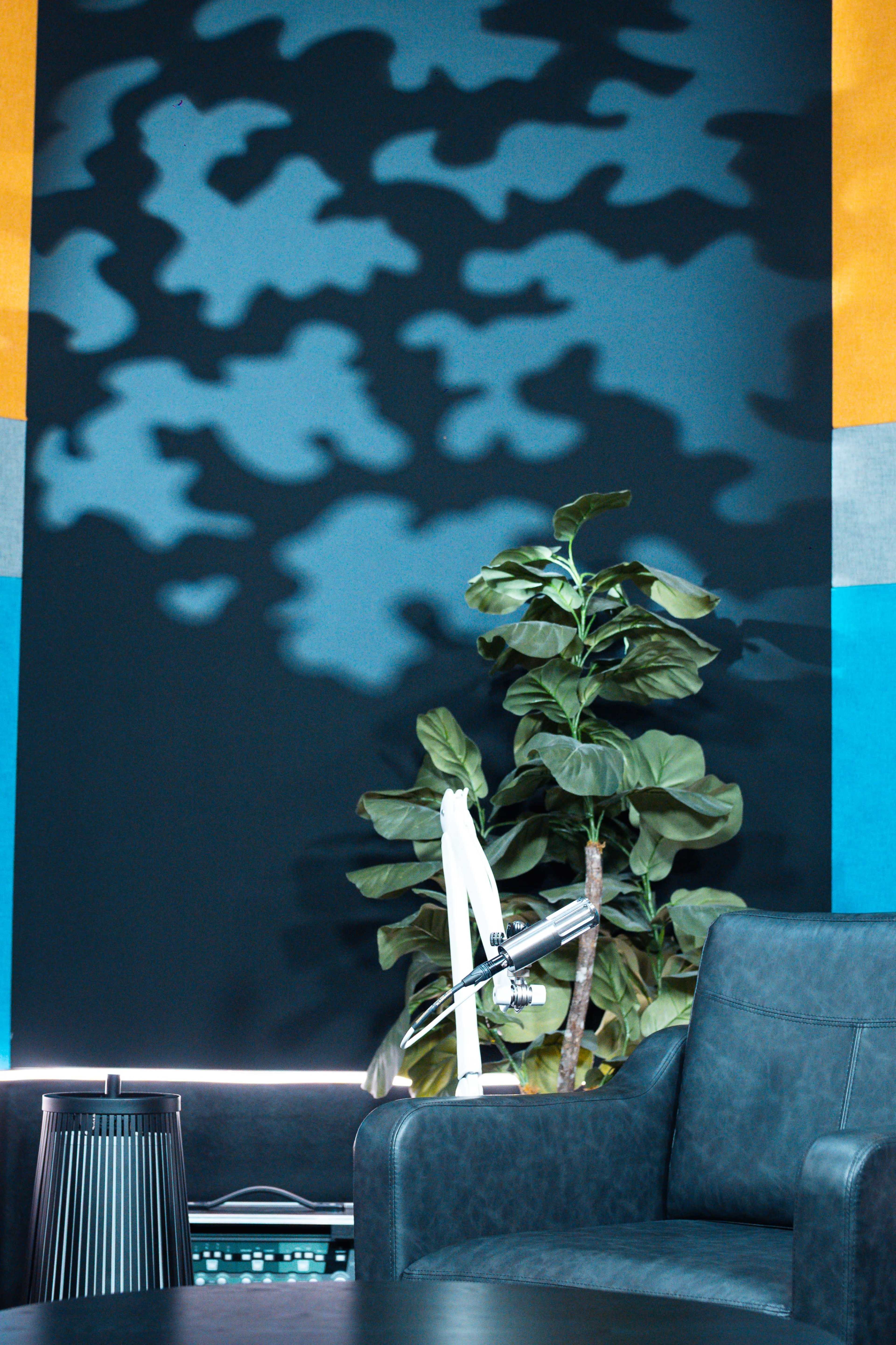 The image shows a corner of a modern room featuring a leafy plant next to a gray armchair, with a large colorful abstract wall in the background.