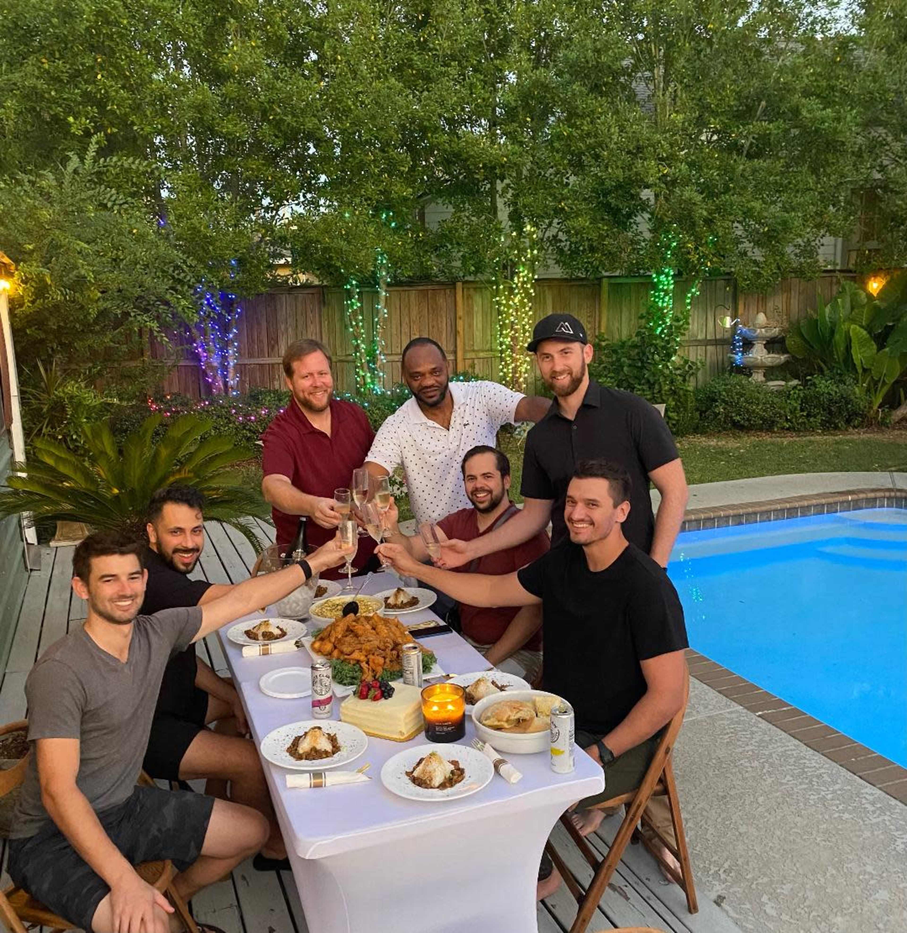 A group of seven men toast with drinks at a table set near a pool, surrounded by greenery and evening lights.