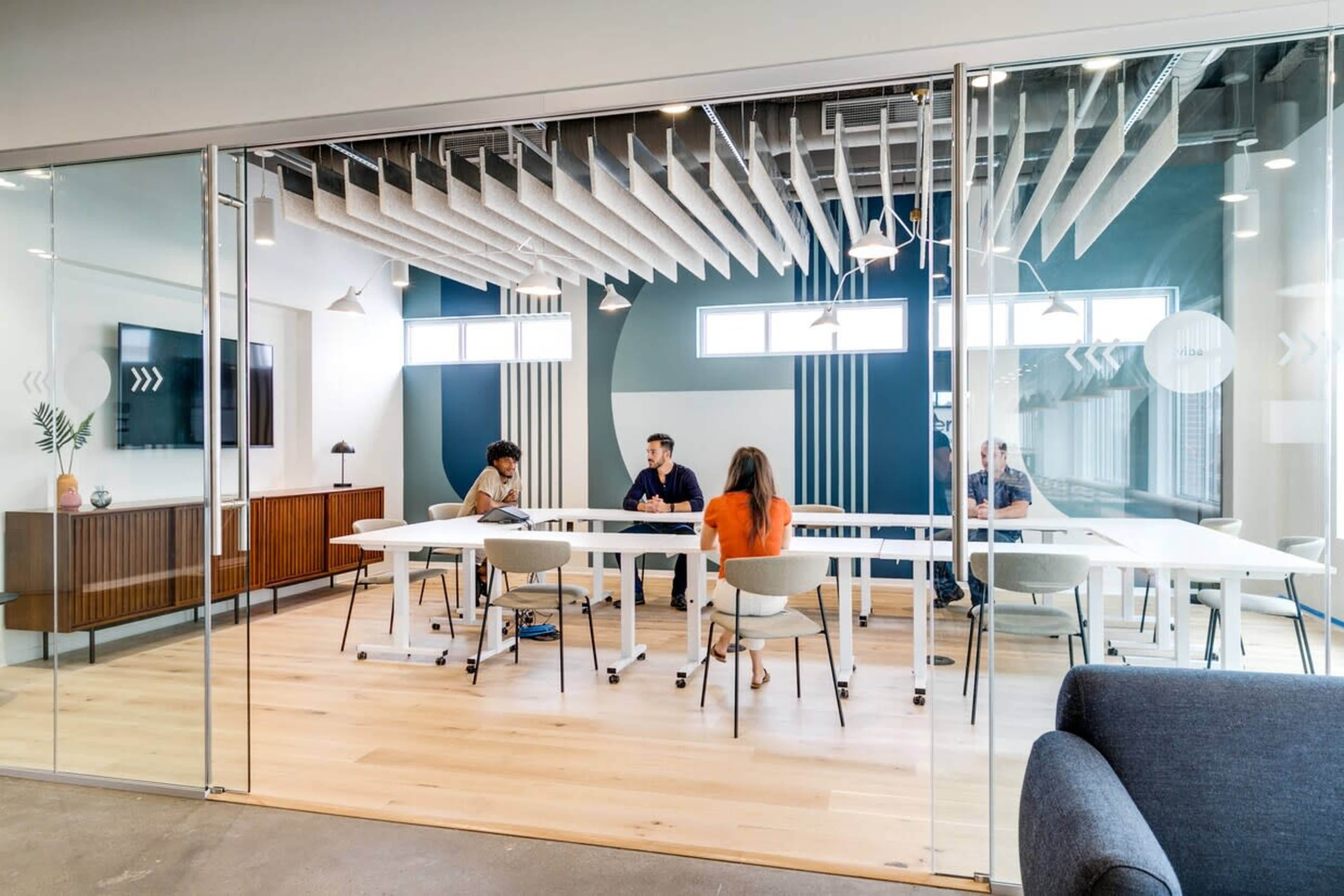 A modern conference room features a long table with four people engaged in discussion, surrounded by glass walls and contemporary design elements.