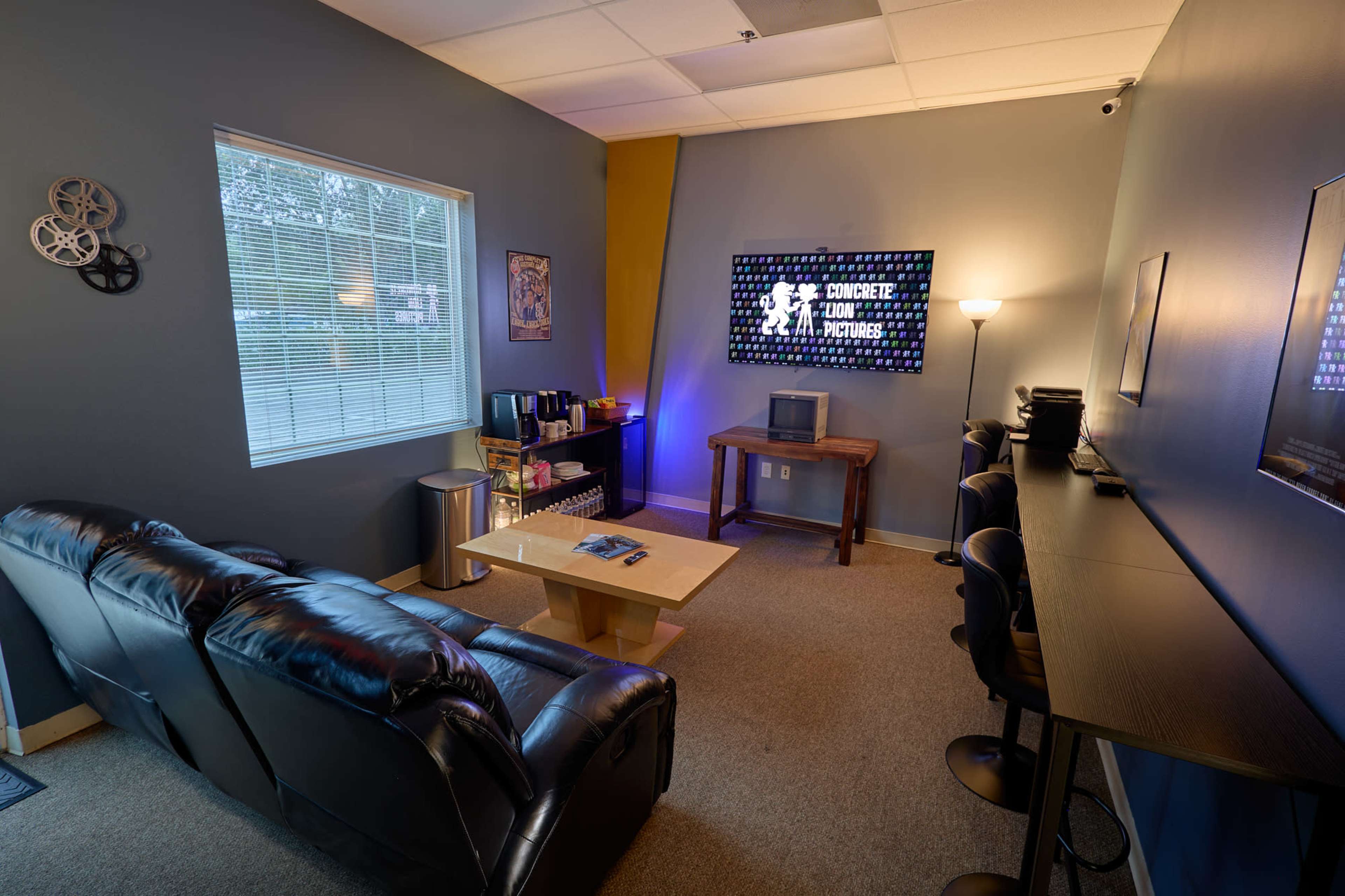 The image shows a small, modern office or studio space with a black leather couch, a wooden table, and a wall-mounted screen displaying "Concrete Pictures."